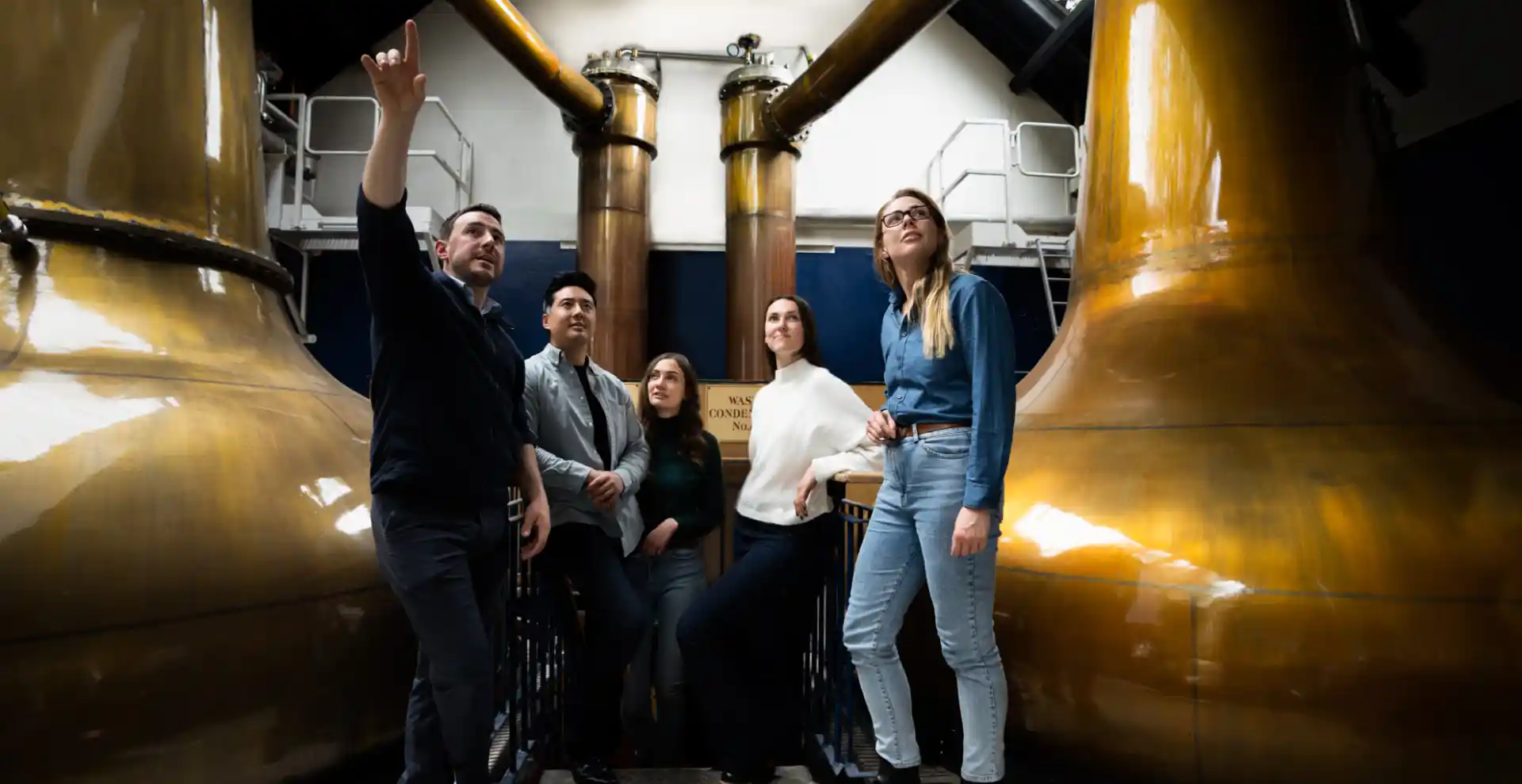 A tour guide shows a group of people round Blair Athol distillery, and points to something behind the camera while standing in front of several whisky stills. 