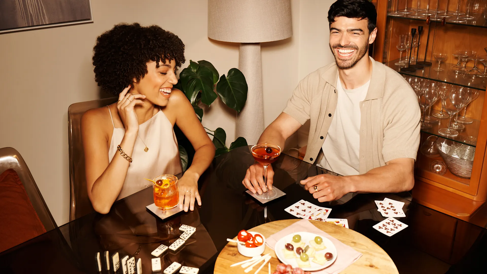 A man and a woman sit at a table drinking cocktails. On the table are dominoes, playing cards and canapes.