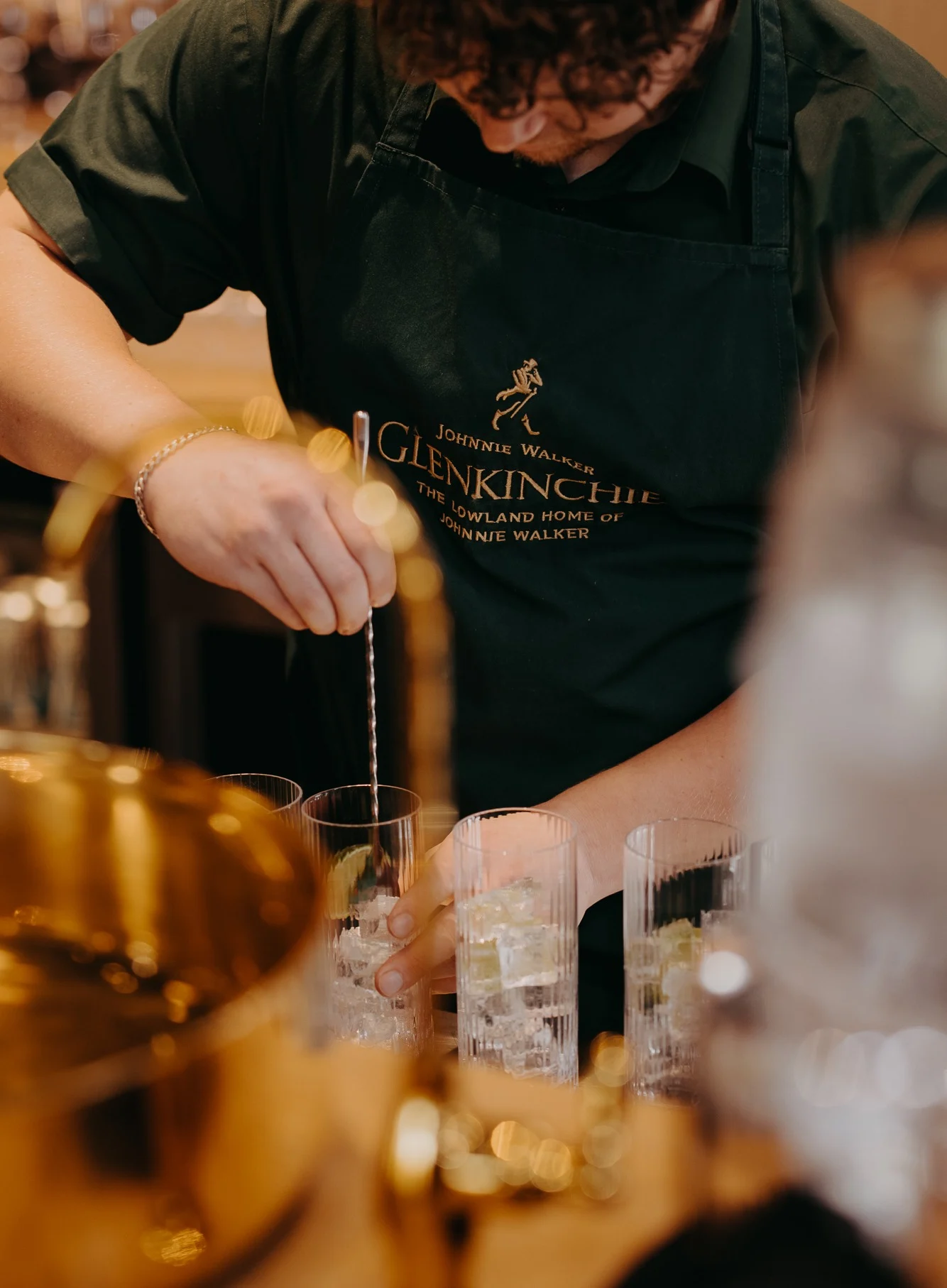 Een barman in een Glenkinchie-schort roert in highballglazen vol ijs.