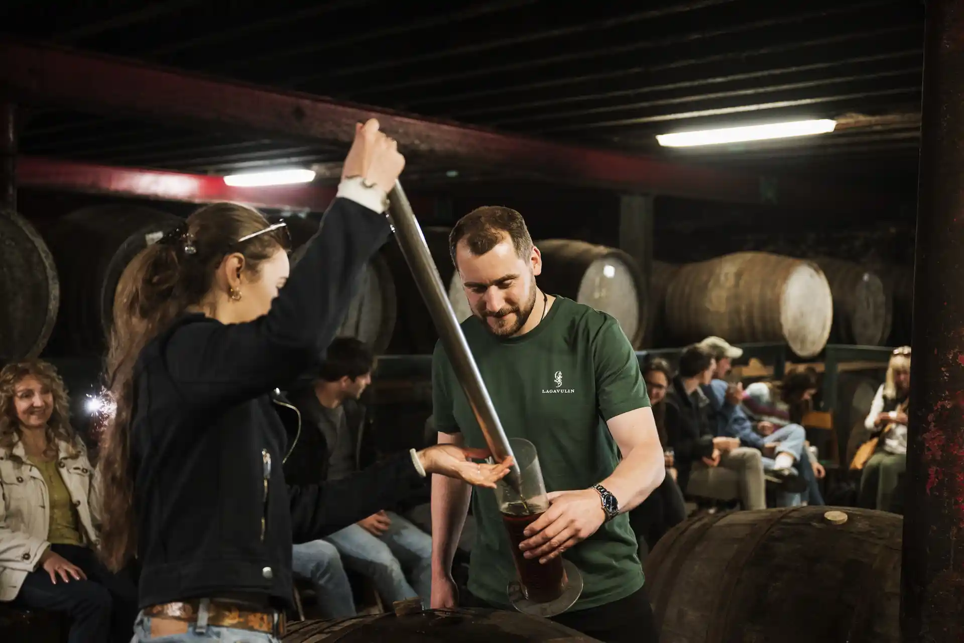 A woman uses a large pipette to take whisky from a wooden barrel and place it into a glass held by a man wearing a green Lagavulin t-shirt. Behind them sits several people and lots of other wooden barrels. 