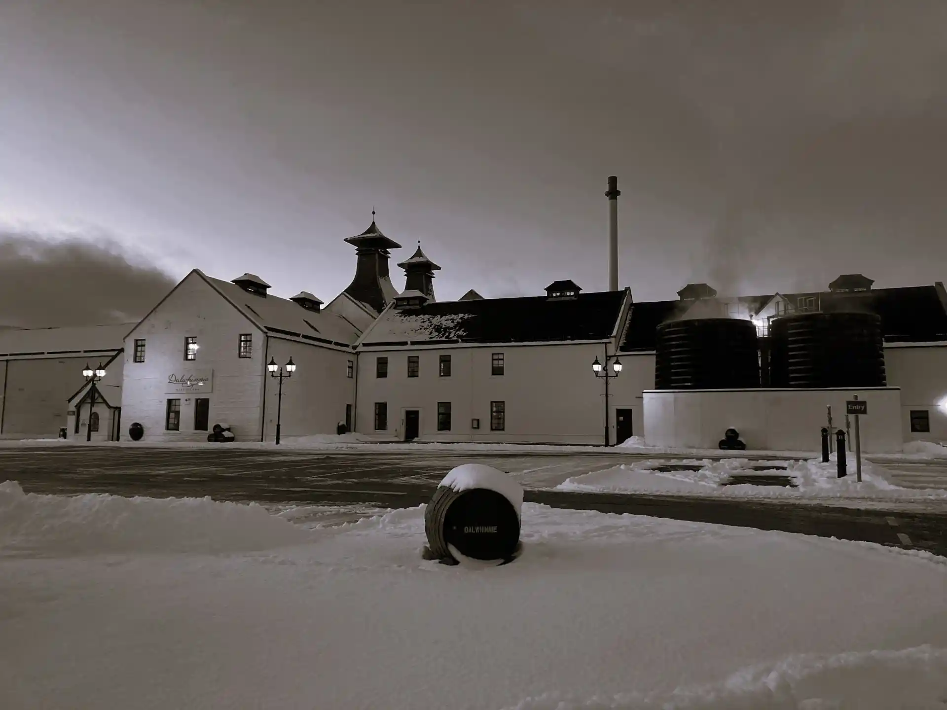 The white buildings of Dalwhinnie distillery are seen from a distance, sitting within the snow. Behind the distillery is a mountain range featuring green trees and a snow-covered summit.