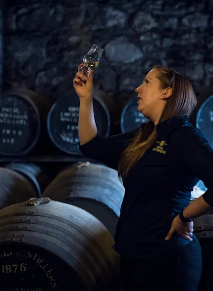 Een vrouw houdt een dramglas met whisky omhoog en inspecteert het, terwijl ze voor een heleboel houten vaten staat.