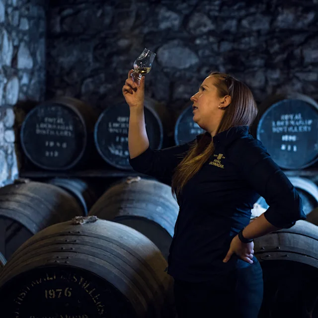 Ontdek meer whisky lingo Een vrouw houdt een dramglas met whisky omhoog en inspecteert het, terwijl ze voor een heleboel houten vaten staat.