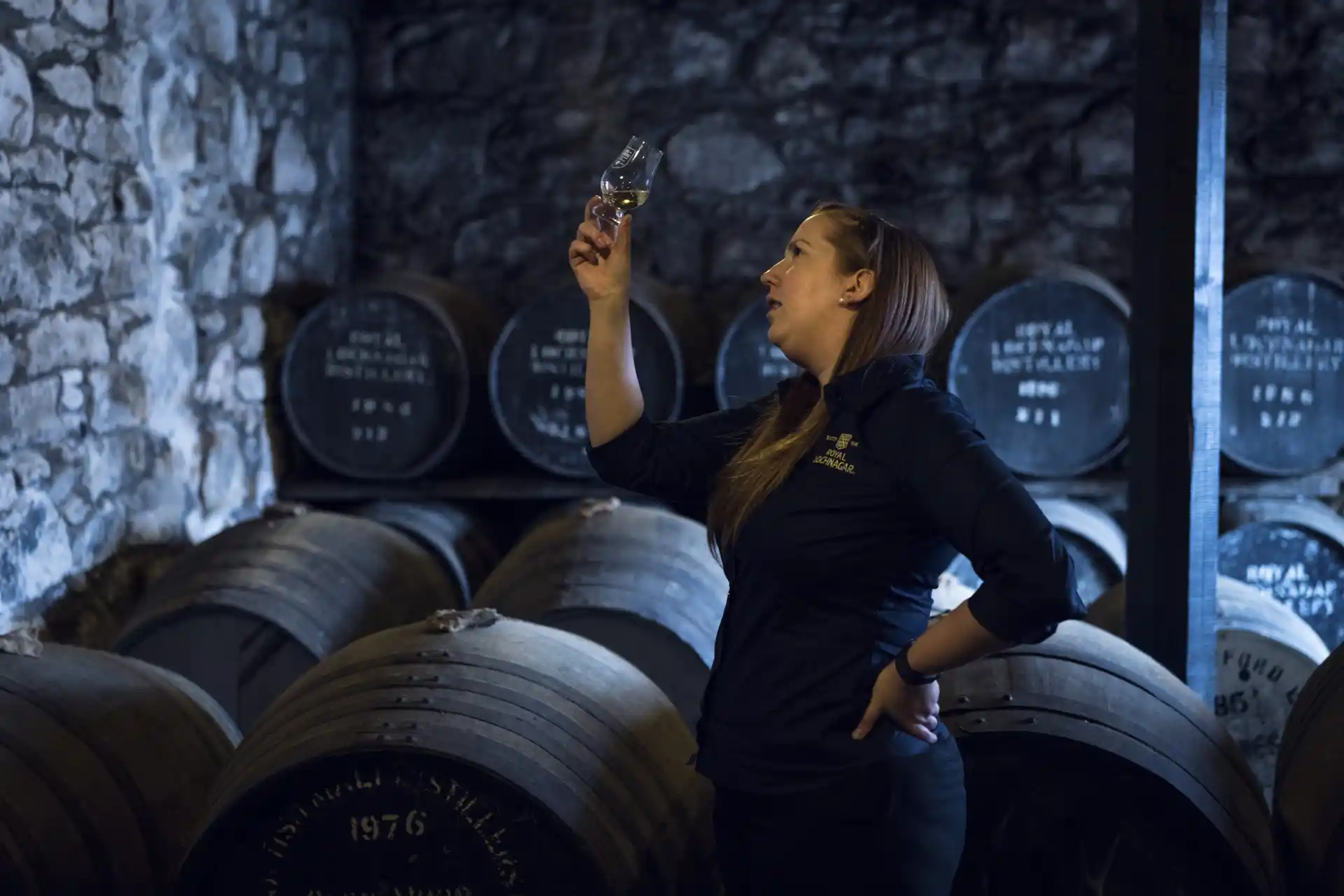 A Royal Lochnagar employee stands in front of several wooden barrels laying on the sides, stacked on shelves. She holds aloft a dram glass containing whisky and looks closely at the glass.