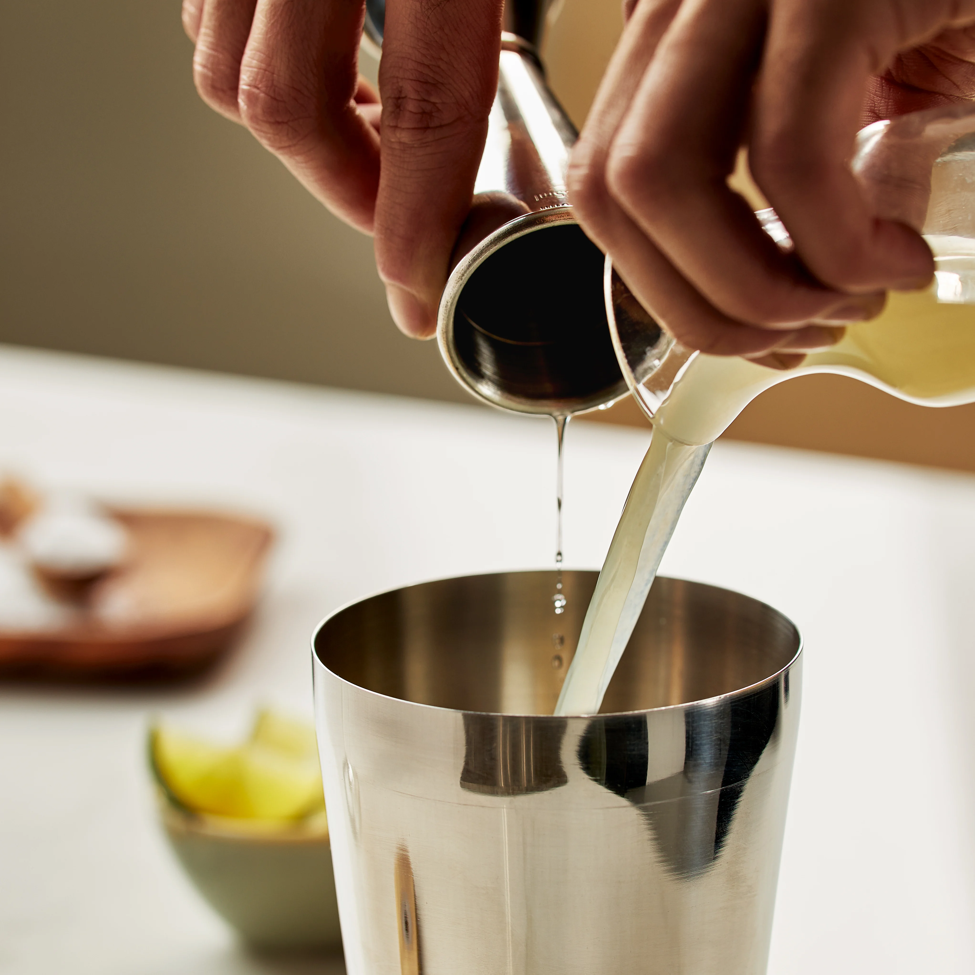 Hands pour tequila from a jigger and lime juice from a cup into a metal cocktail shaker.  