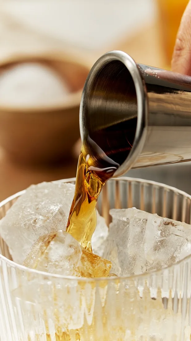 Een donkere gedistilleerde drank wordt uit een zilveren jigger in een met ijs gevuld glas gegoten.