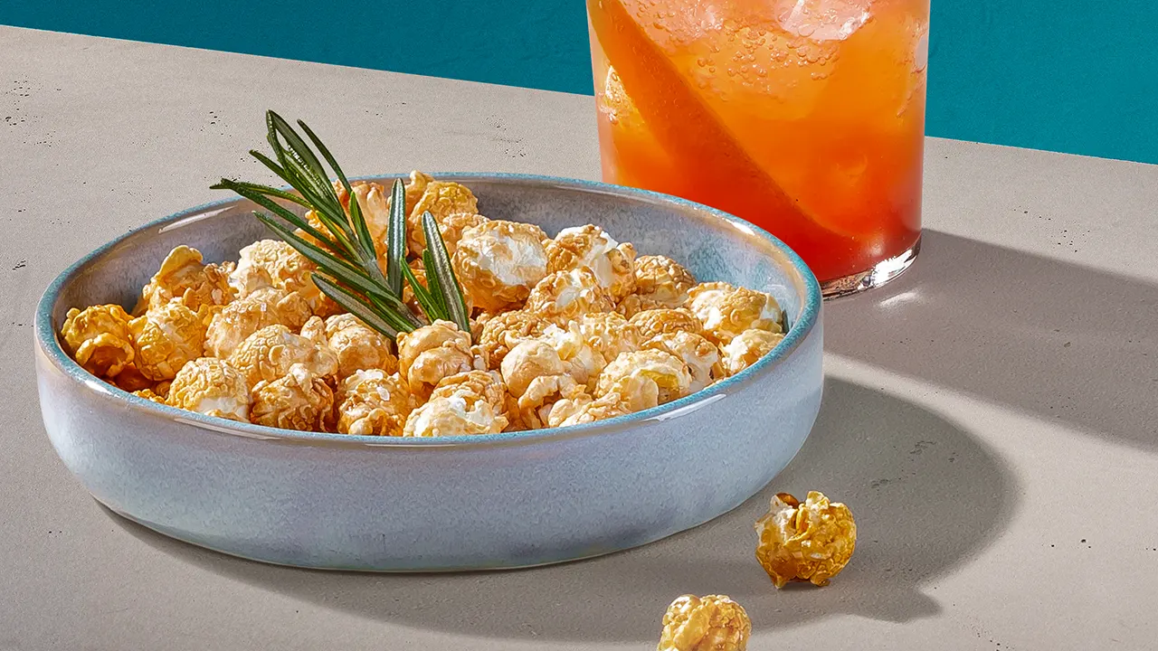 An orange-coloured whisky cocktail next to a bowl of toasted popcorn with sea salt.