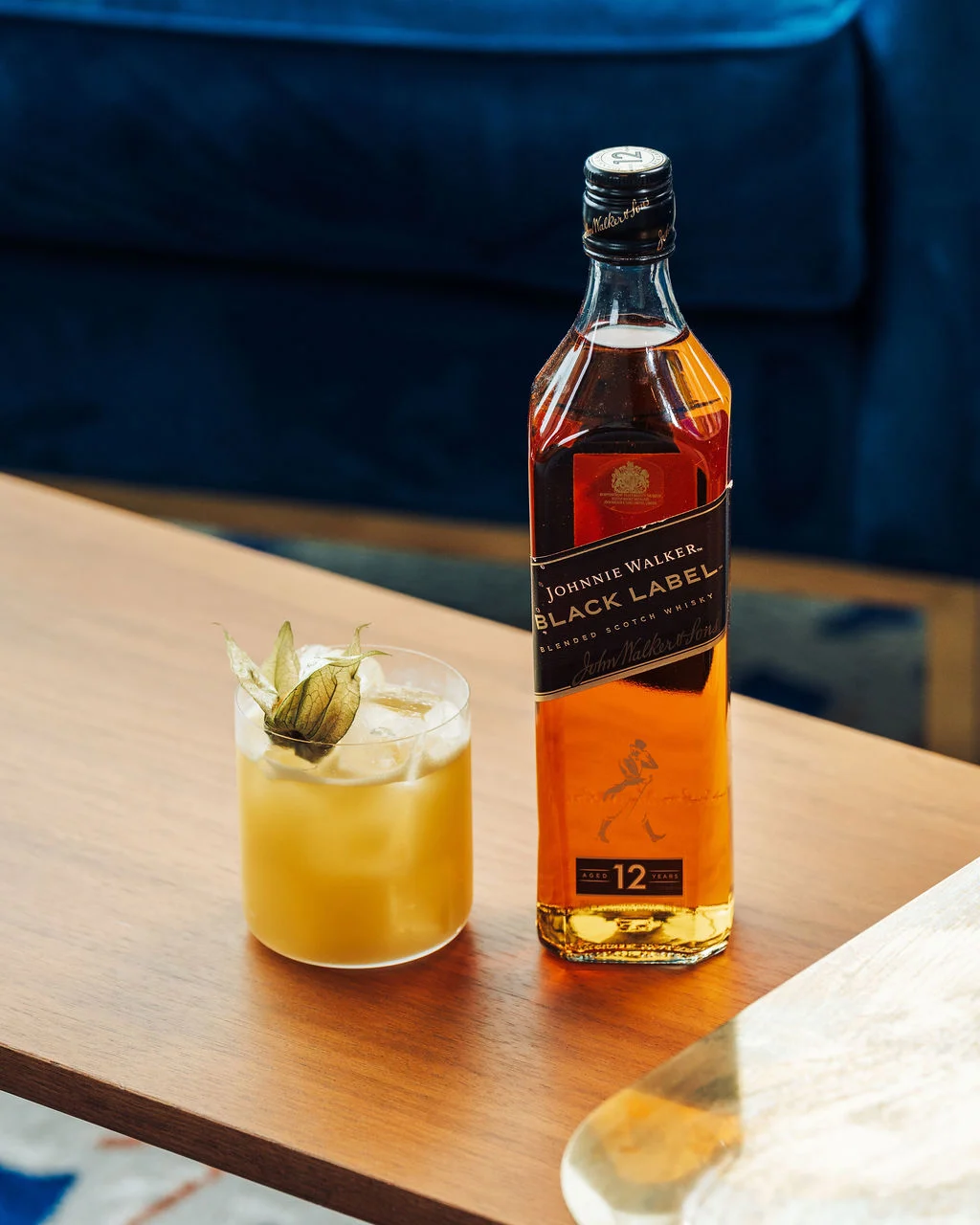 A bottle of Johnnie Walker Black Label on a wooden table next to a light yellow cocktail in a rocks glass with a green herb garnish