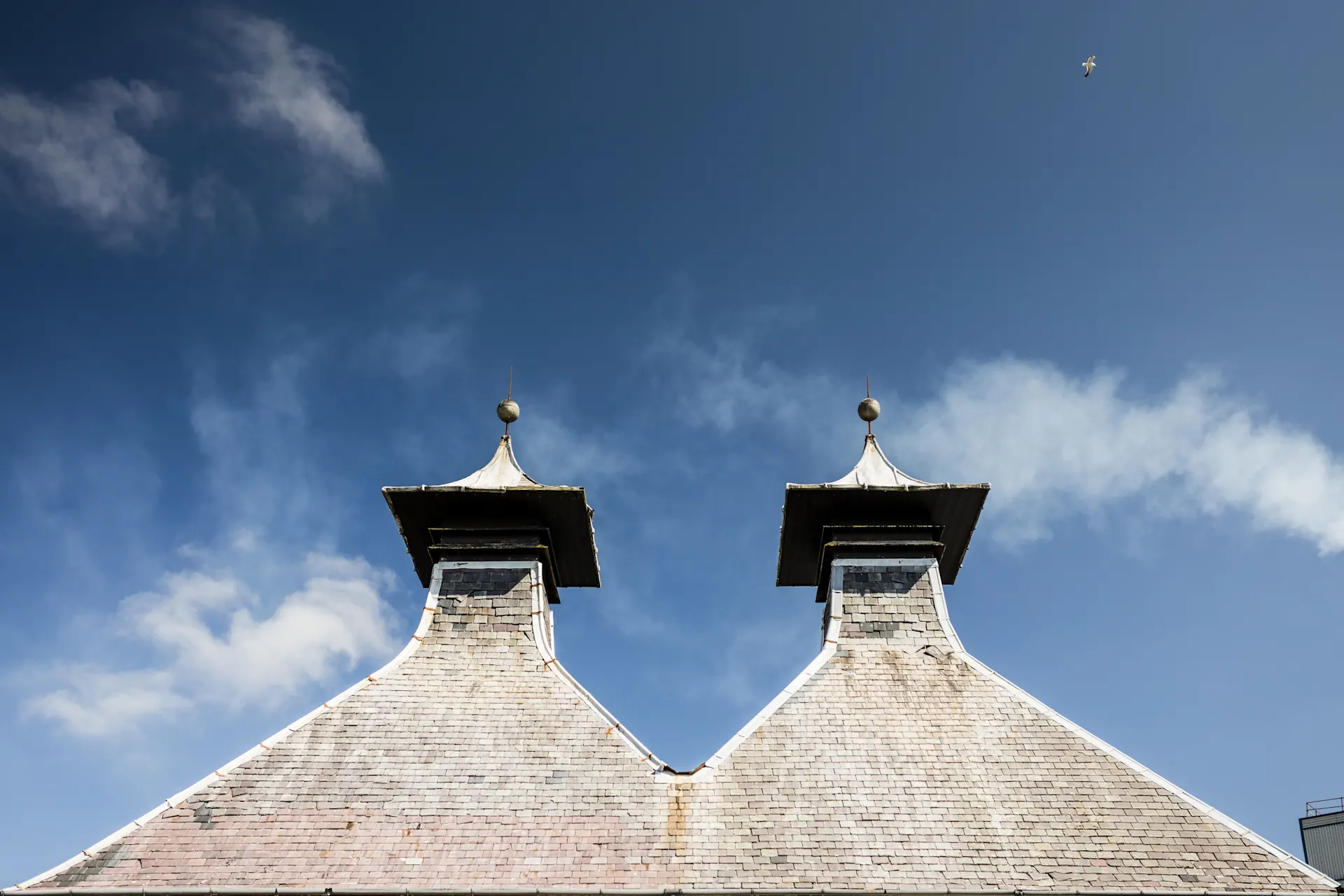 Two pitched roofs of Port Ellen distillery are set against a bright blue sky.