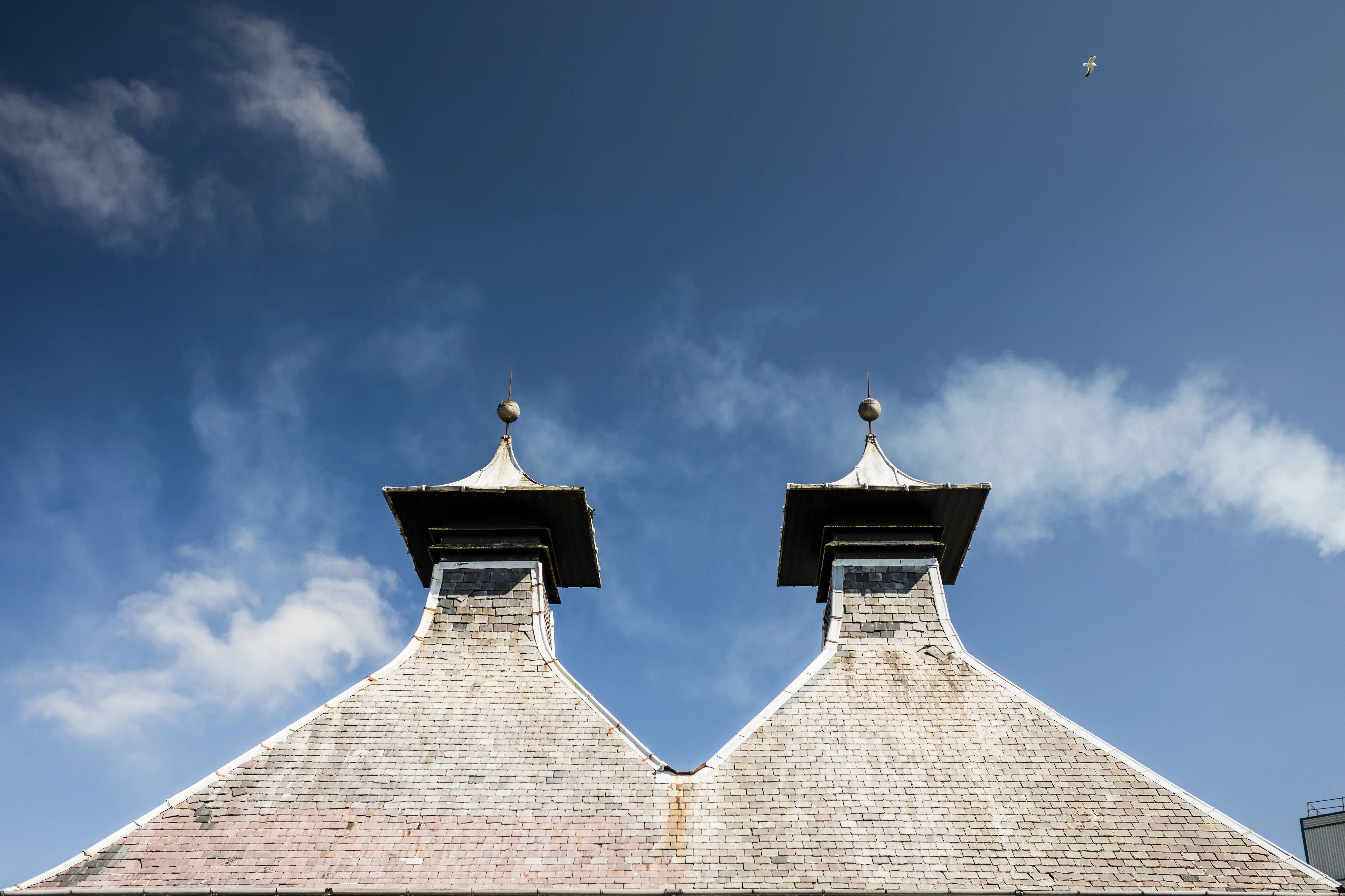 Two pitched roofs of Port Ellen distillery are set against a bright blue sky.