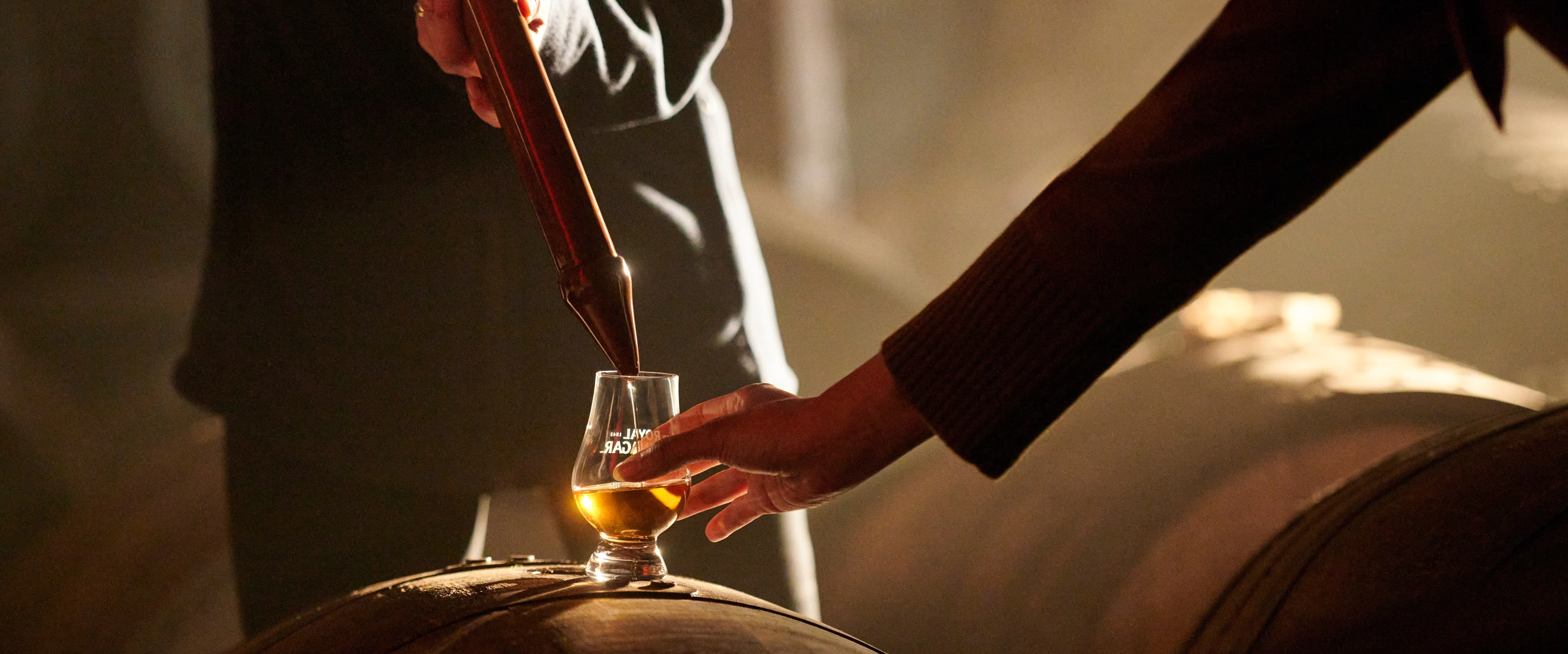 A distillery worker pouring a sample of Scotch whisky from a valinch into a Glencairn glass resting on a wooden cask during a warehouse tasting experience