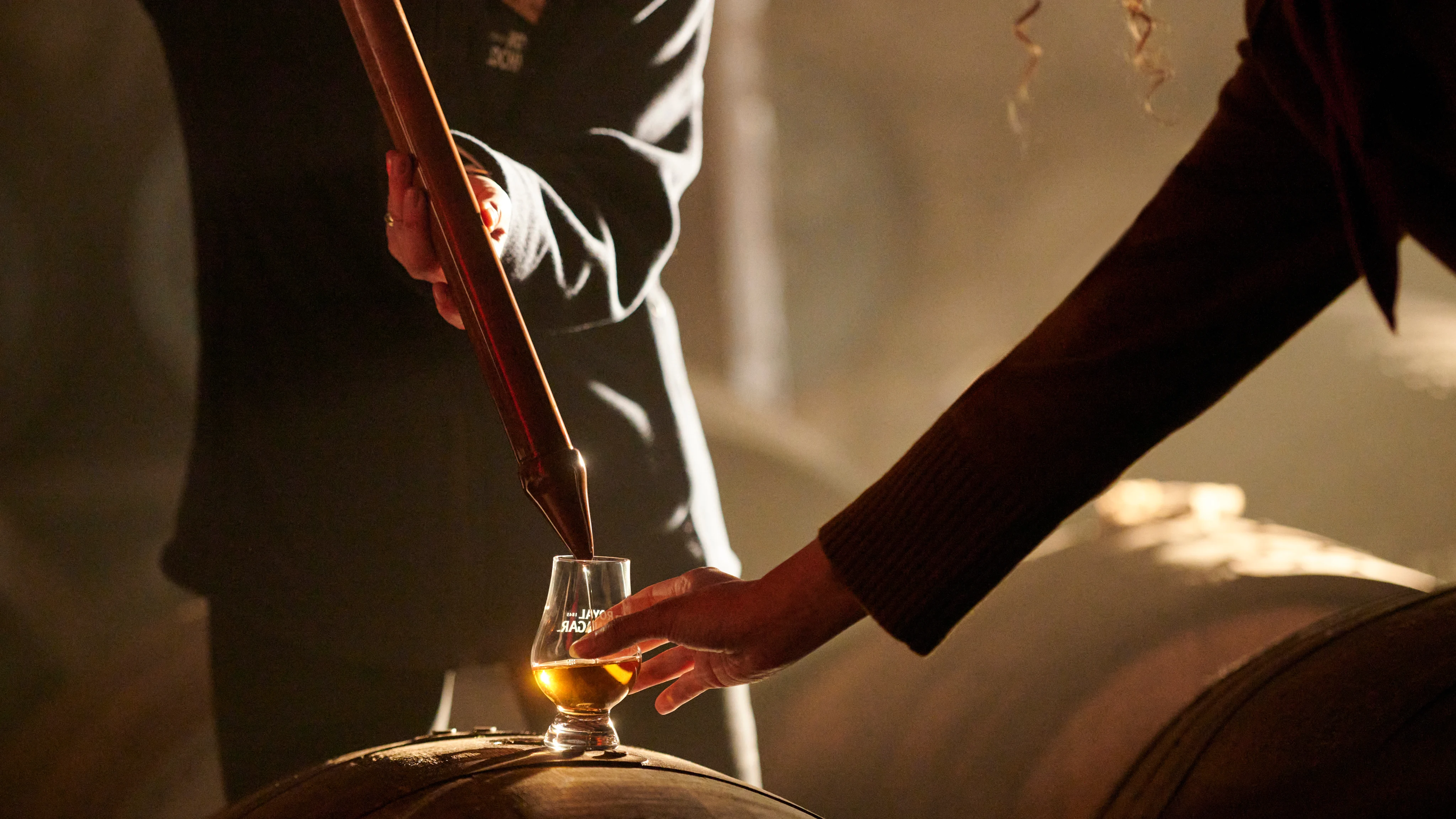 A distillery worker pouring a sample of Scotch whisky from a valinch into a Glencairn glass resting on a wooden cask during a warehouse tasting experience