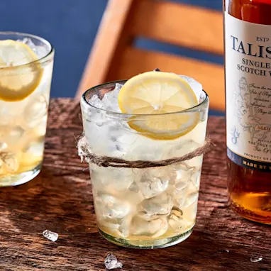 Two lowball whisky cocktails sit on a wooden board next to a bottle of Talisker 10 year old single malt whisky