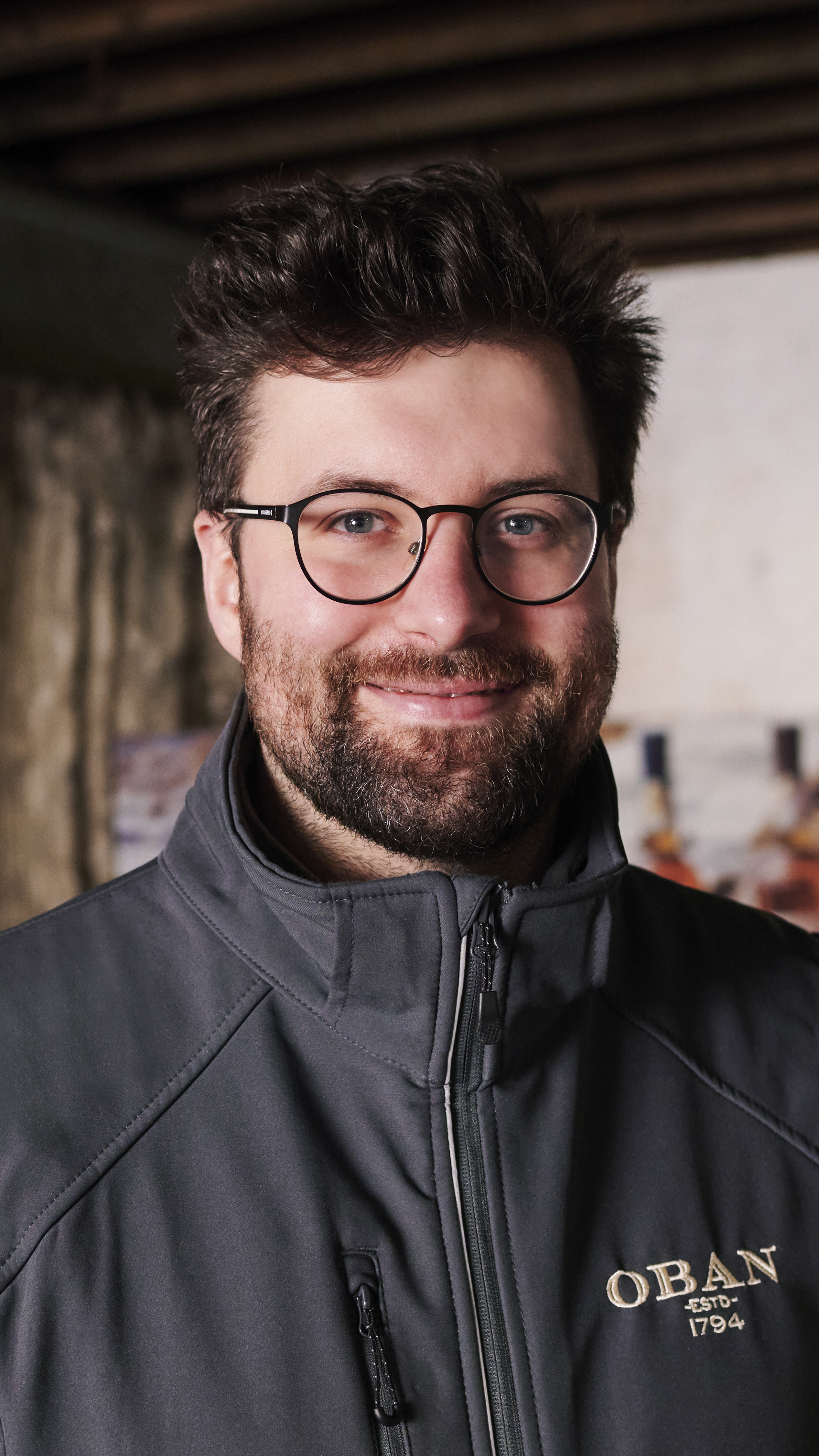 Callum, een reisleider van Oban, staat op de foto in een opslagruimte van de distilleerderij, met een grijs Oban-jasje aan.