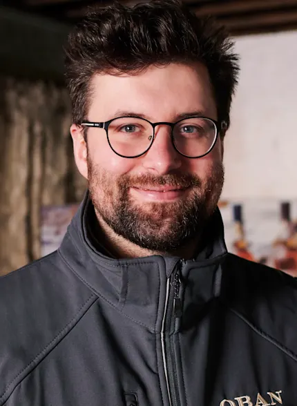 Callum, ein Oban-Reiseleiter, steht in einem Lagerraum der Brennerei und trägt eine graue Oban-Jacke.