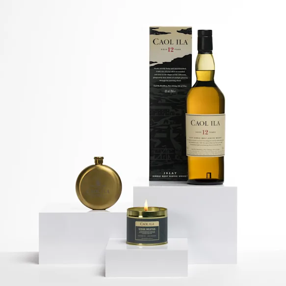 Caol Ila 12 Year Old whisky bottle with a matching box, a gold flask, and a lit whisky-scented candle on white display blocks.