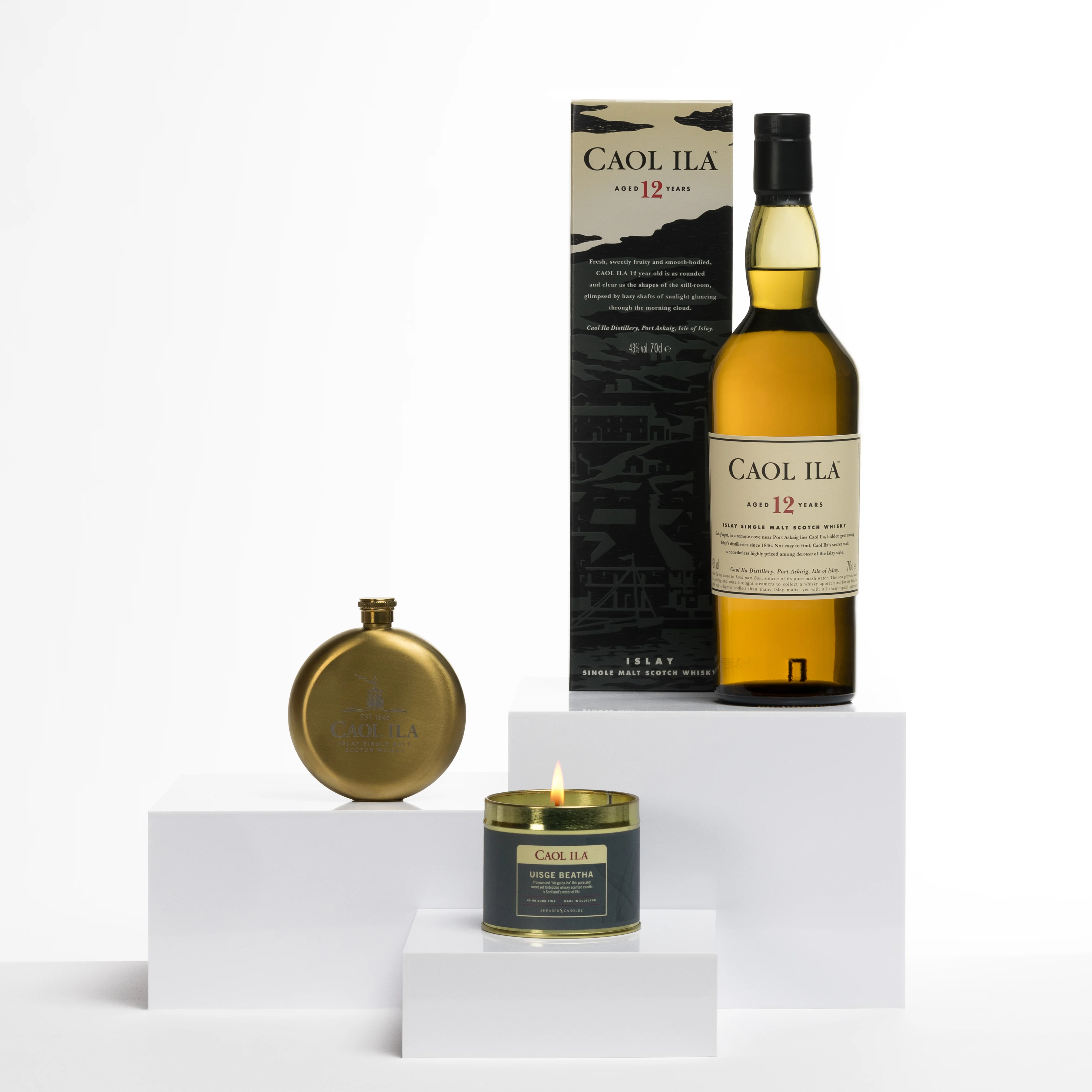 Caol Ila 12 Year Old whisky bottle with a matching box, a gold flask, and a lit whisky-scented candle on white display blocks.