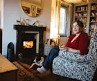 Caroline McQuistin sits in an armchair with her border collie dog in front of a roaring fireplace, holding a rocks glass containing whisky.