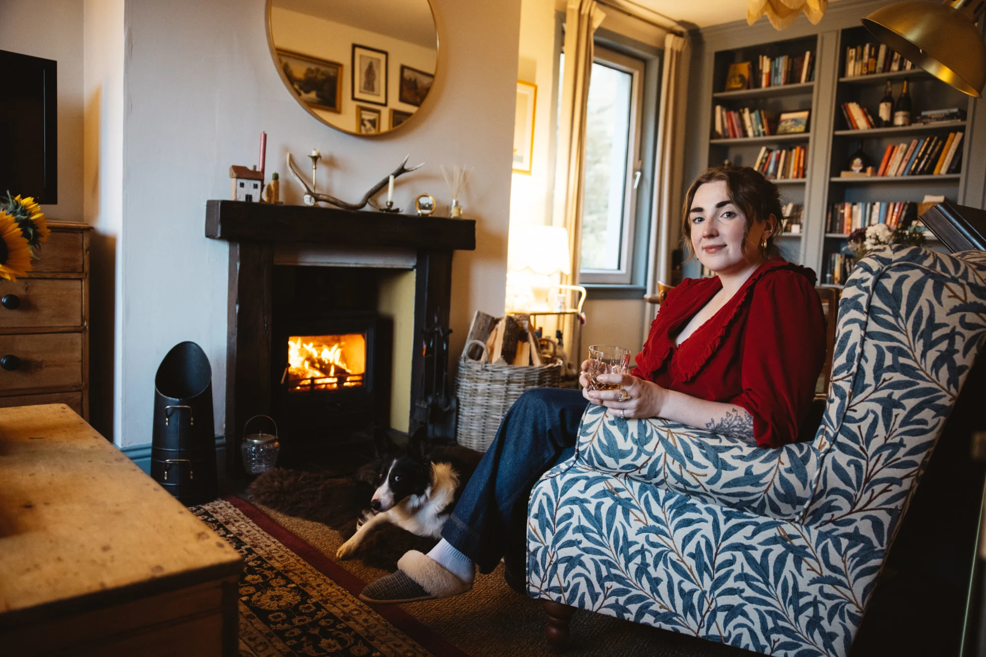Caroline McQuistin sitzt mit ihrem Border-Collie-Hund in einem Sessel vor dem prasselnden Kamin und hält ein Steinglas mit Whisky in der Hand.