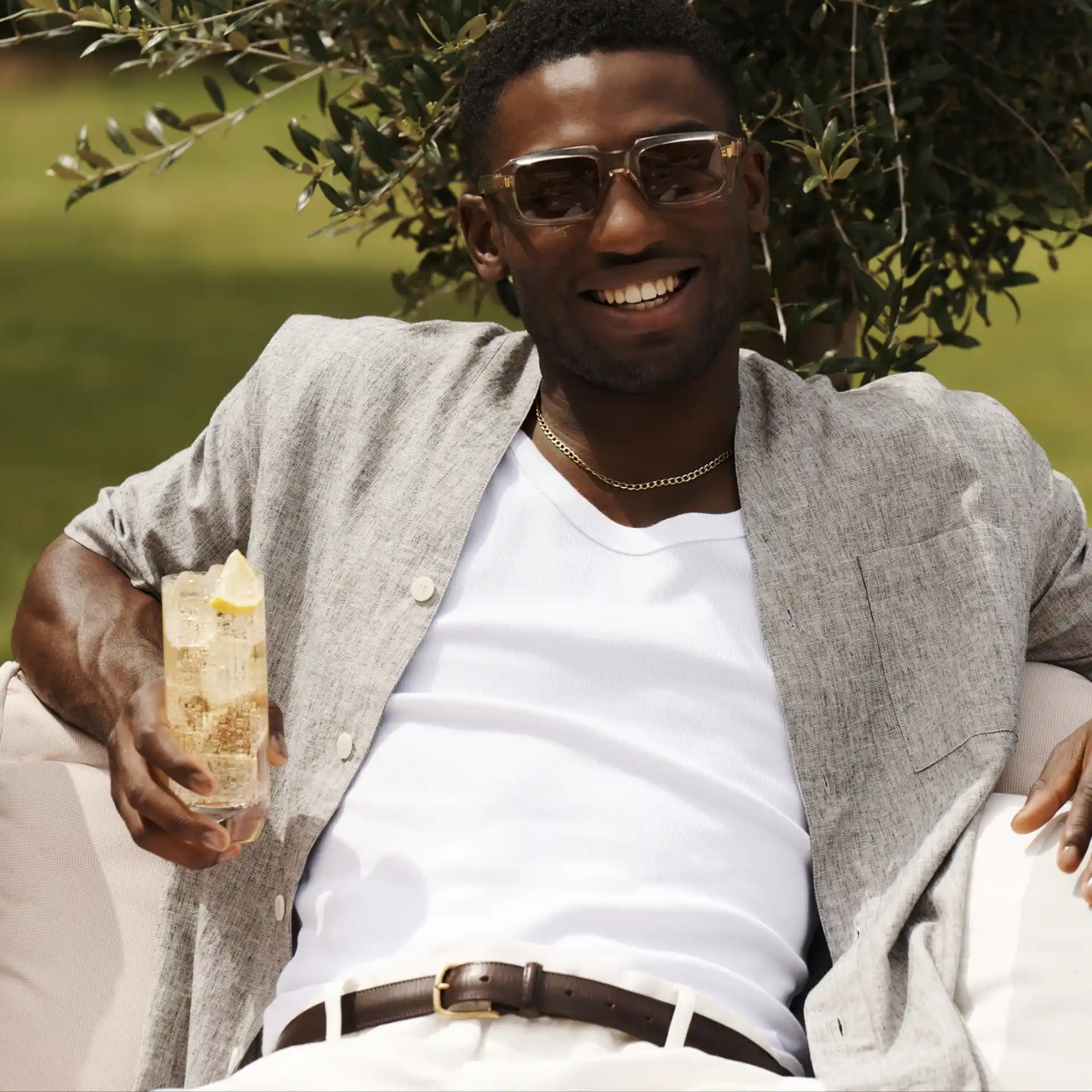 A man sits on a sofa outdoors holding a whisky highball cocktails while smiling.