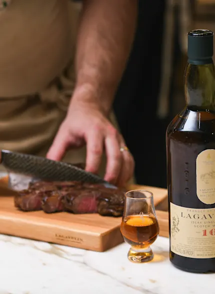 A man cuts slices of steak on a wooden board, next to a bottle of Lagavulin and a dram glass containing whisky.
