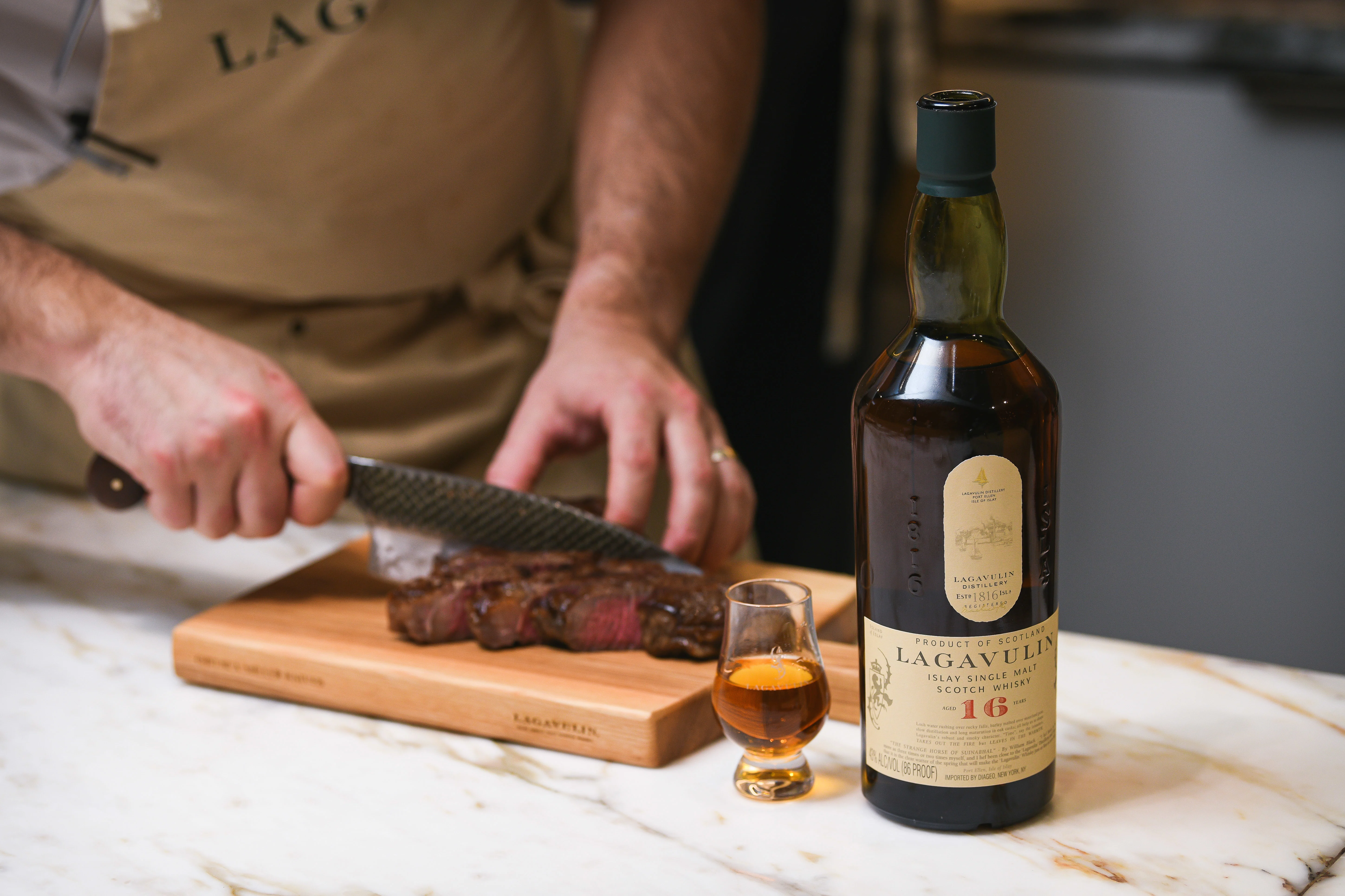 A man cuts slices of steak on a wooden board, next to a bottle of Lagavulin and a dram glass containing whisky.