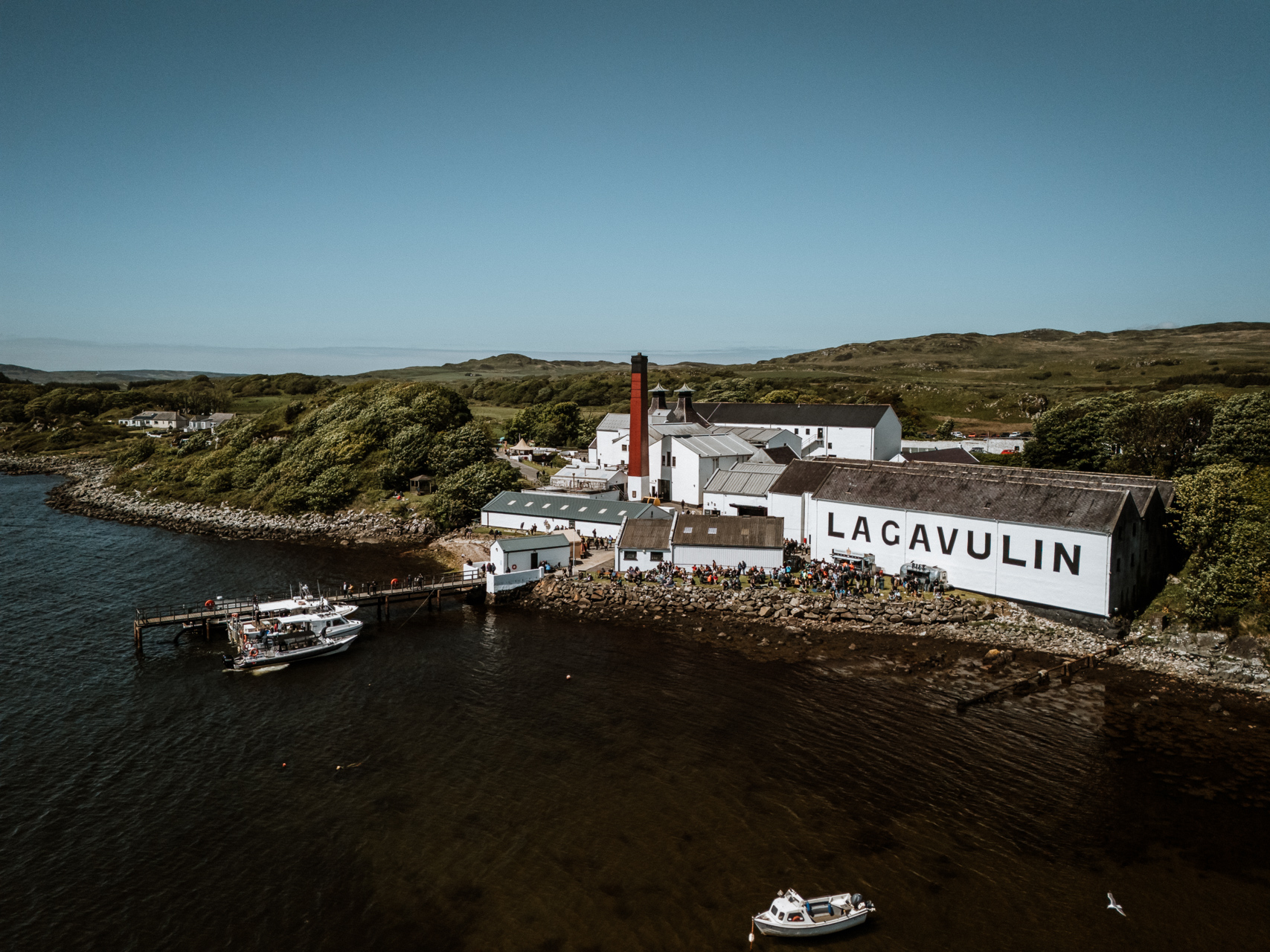 Die Lagavulin-Destillerie mit blauem Himmel und den dahinter liegenden Küsten und Hügeln.