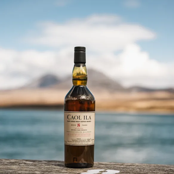 Caol Ila 8 Year Old Fèis Ìle 2025 Edition bottle standing on a wooden surface with the sea and distant hills in the background.