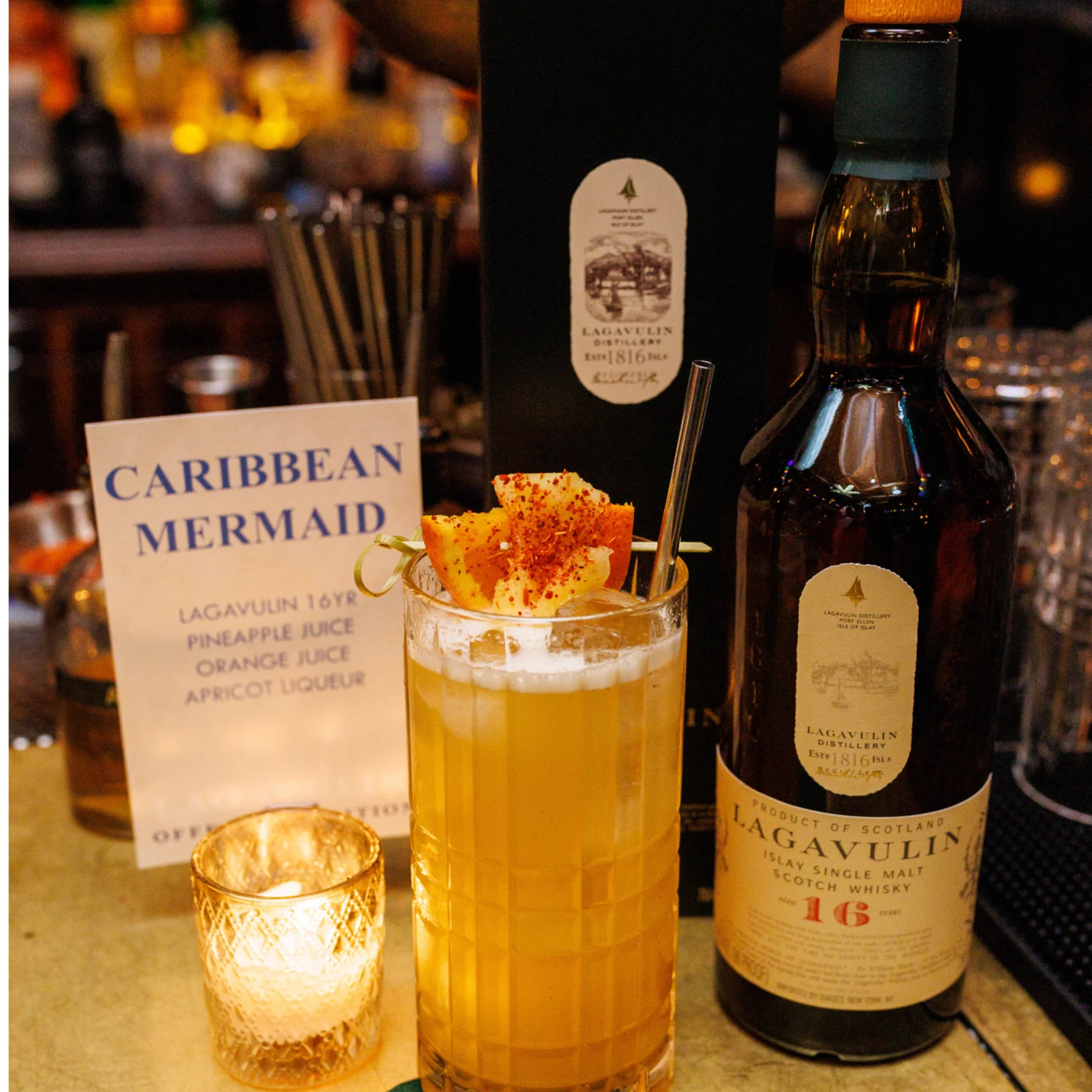 Orange drink garnished with an orange wedge next to a menu saying 'Caribbean Mermaid' and a bottle of Lagavulin whisky