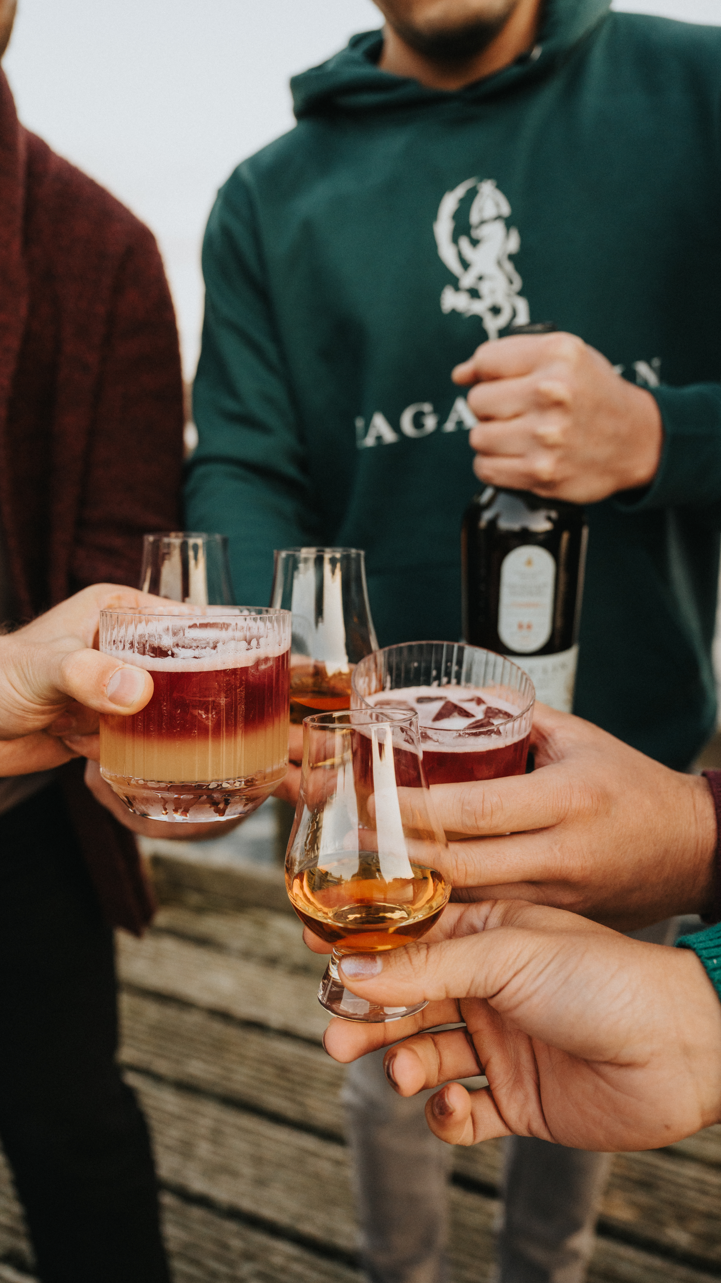 Five people clinking glasses with cocktails and whiskey; one person holds a bottle. The group stands outdoors on wooden decking.