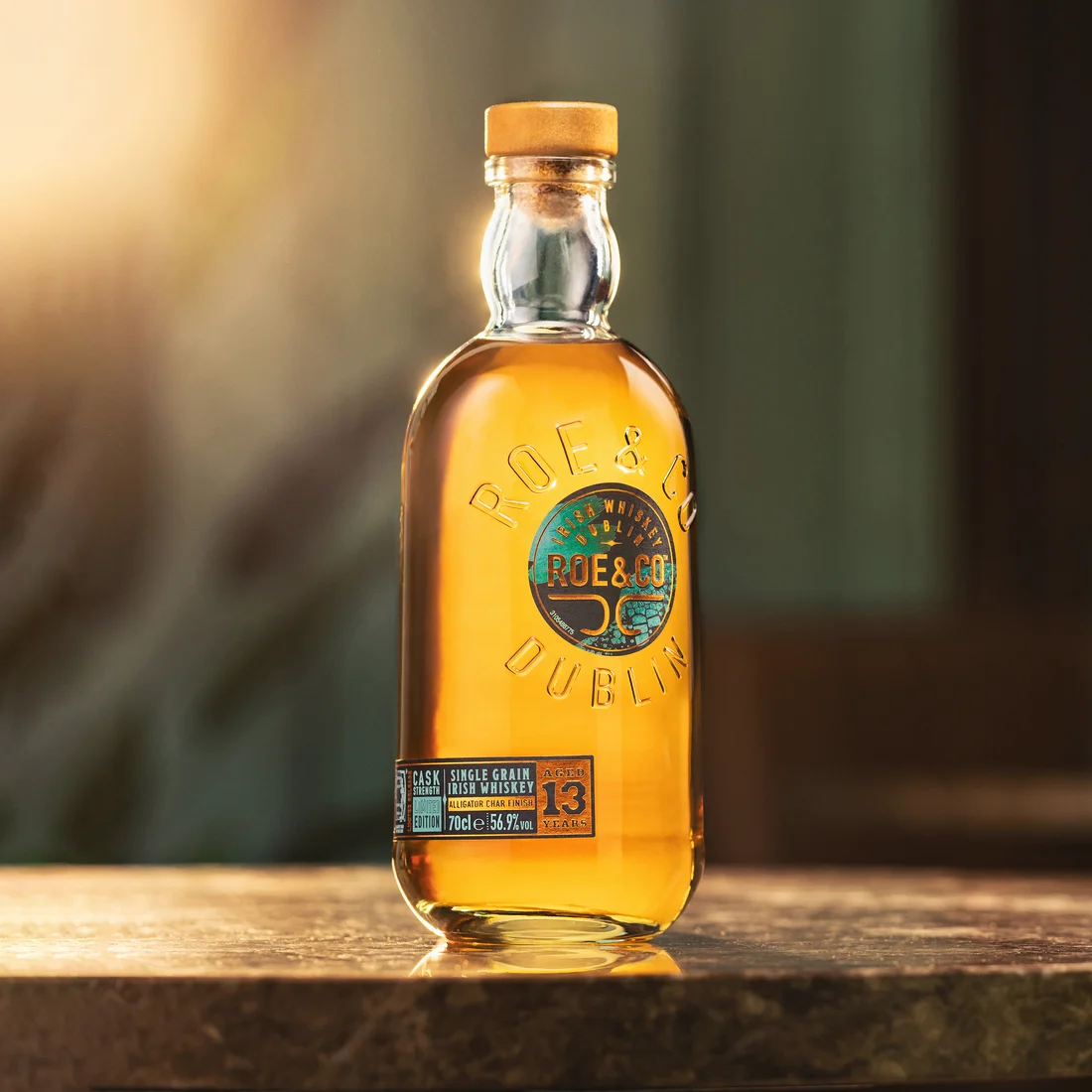 Die Flasche Roe & Co 13-Year-Old Cask Strength Irish Whiskey steht allein auf einem Tisch, in weiches Sonnenlicht getaucht, mit einem unscharfen Hintergrund.