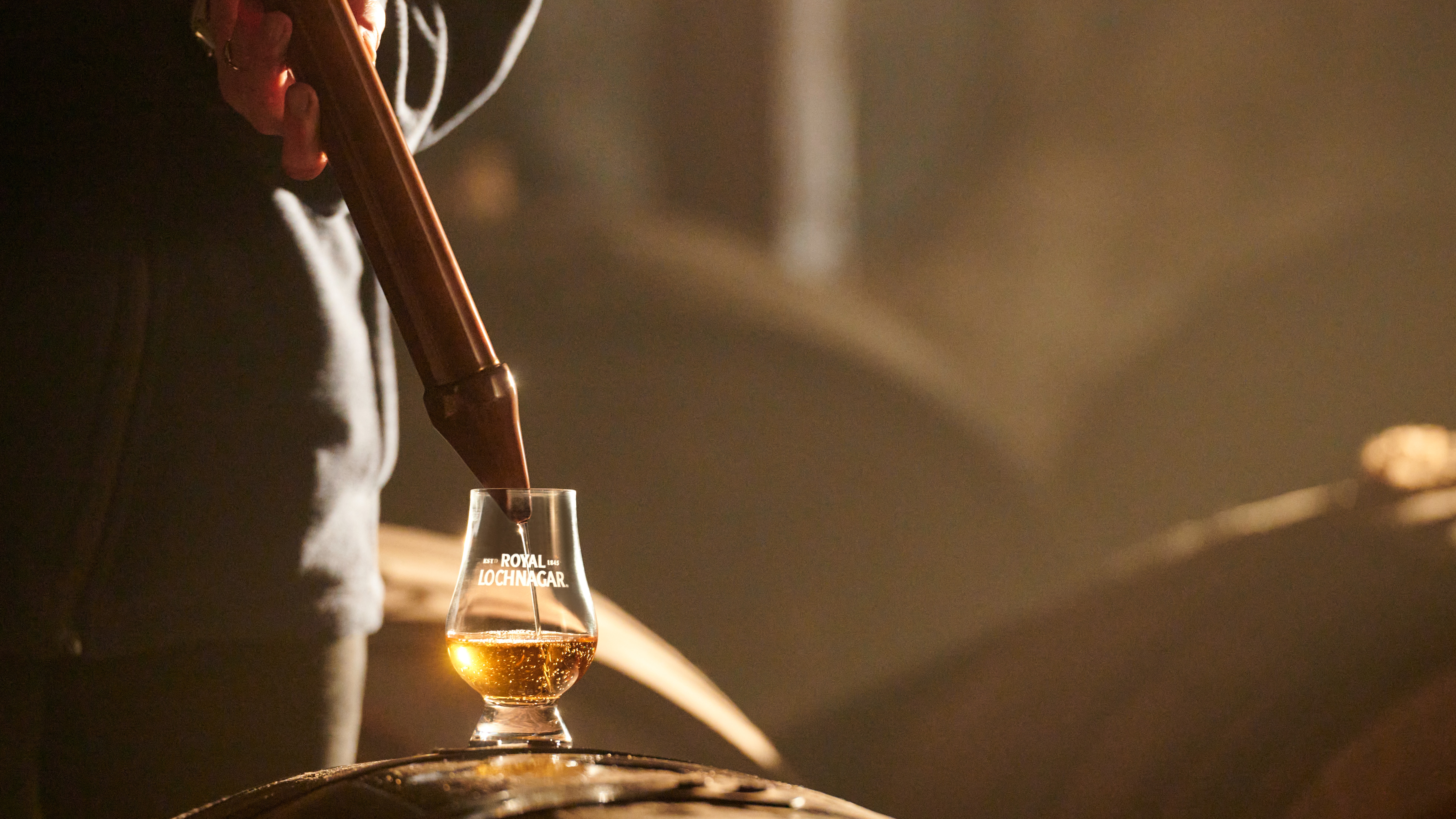 Ein kleines Glas mit Whisky steht auf einem Holzfass. Das Glas wird mit einer großen Metallpipette befüllt. Auf dem Glas ist der Schriftzug "Royal Lochnagar" aufgedruckt.