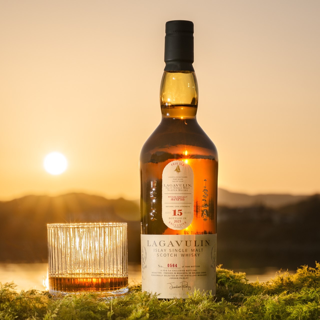 Lagavulin 15 Year Old Fèis Ìle 2025 Edition Flasche und ein Whiskyglas auf Moos gestellt mit dem Sonnenuntergang im Hintergrund.