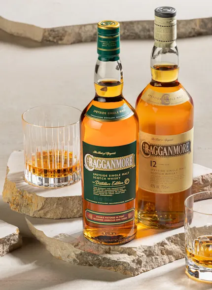 Two bottles of Cragganmore whisky sit on rough neutral marble slabs. Either side of the bottles are two rocks glasses containing whisky.