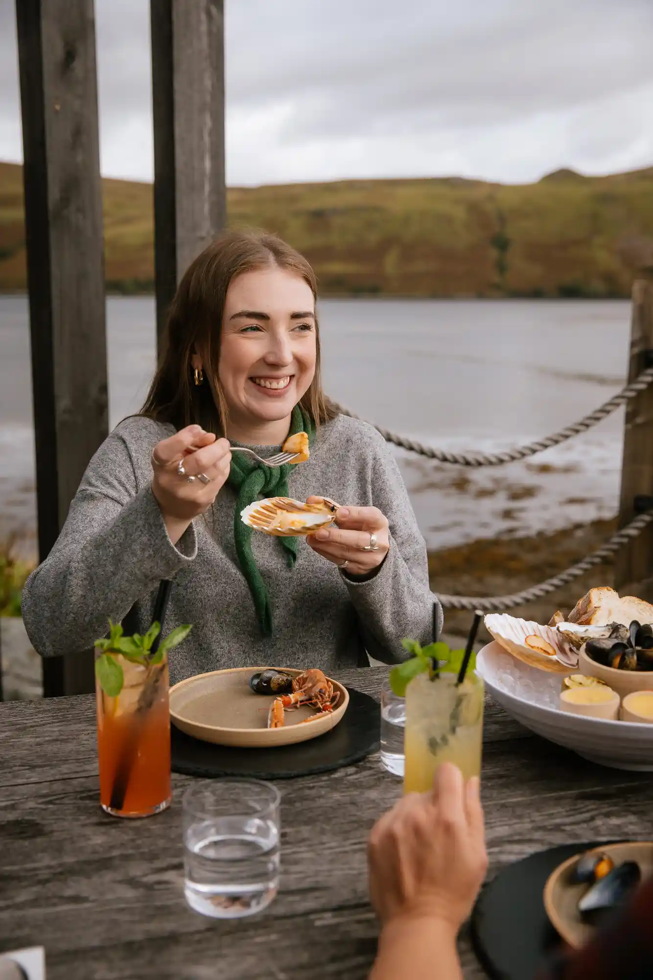 Influencer Caroline McQuistin at Talisker distillery, sitting at a wooden table outside laden with seafood and whisky cocktails