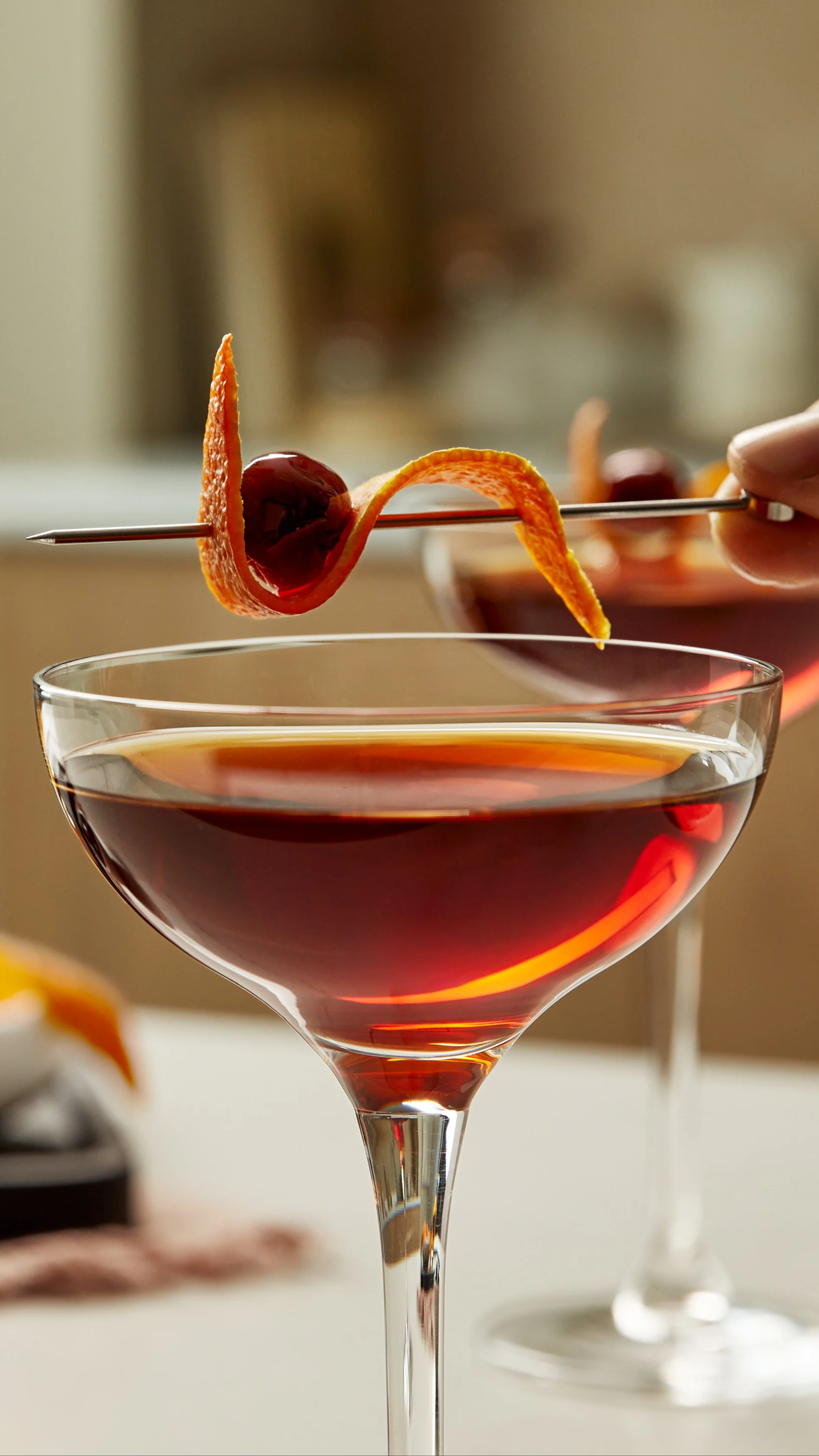 Eine Person legt eine aufgespießte Orangenschale und eine Kirsche auf den Rand eines braunen Cocktails in einem Coupé-Glas