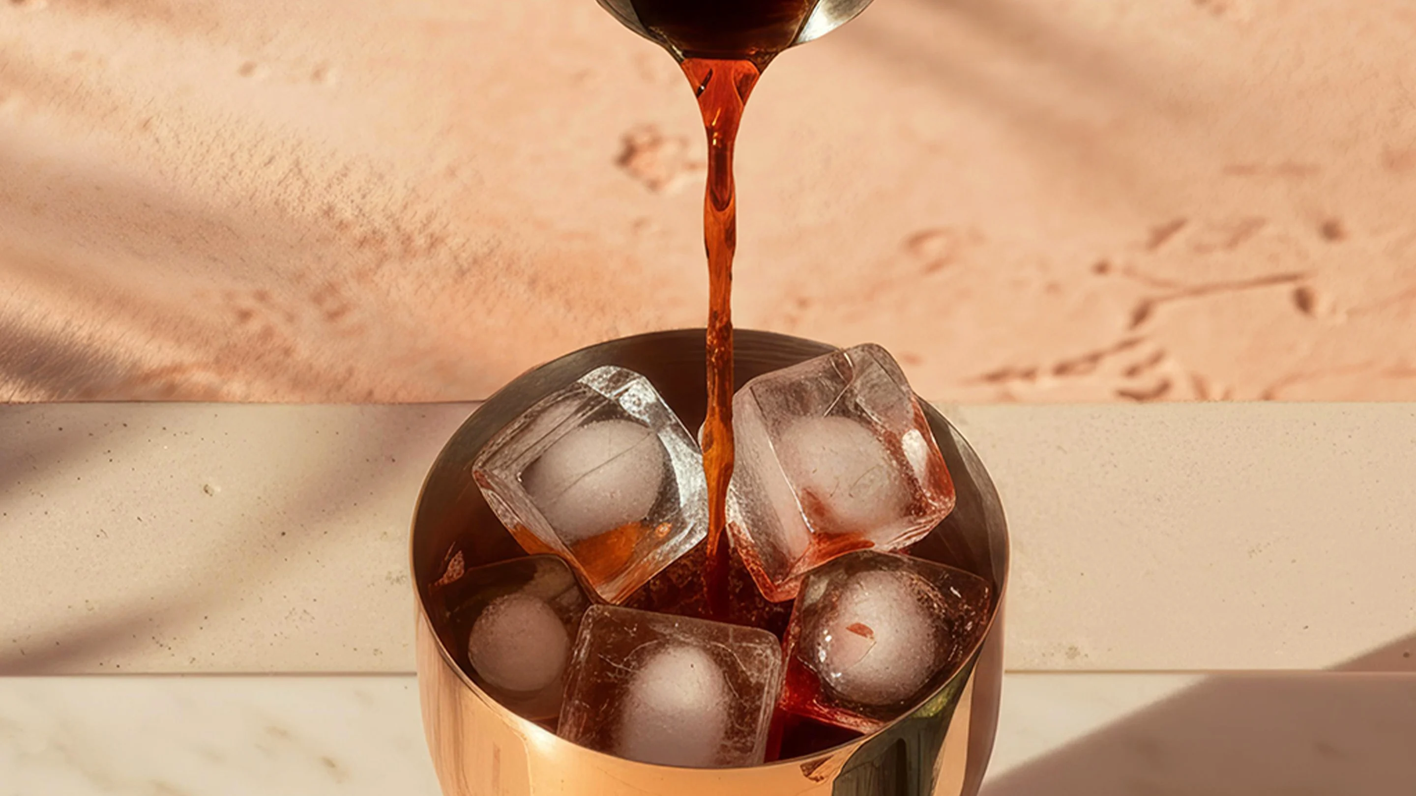A brown liquid is poured into a copper cocktail shaker over ice, with a bowl of green olives nearby