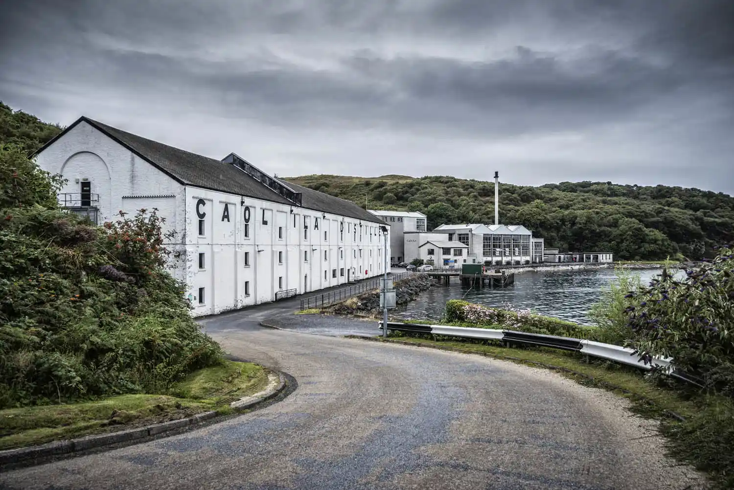 Caol Ila distillery is seen from a winding road which leads to the front of the building where the distillery meets the bay. Behind the distillery are hills with lots of green foliage.