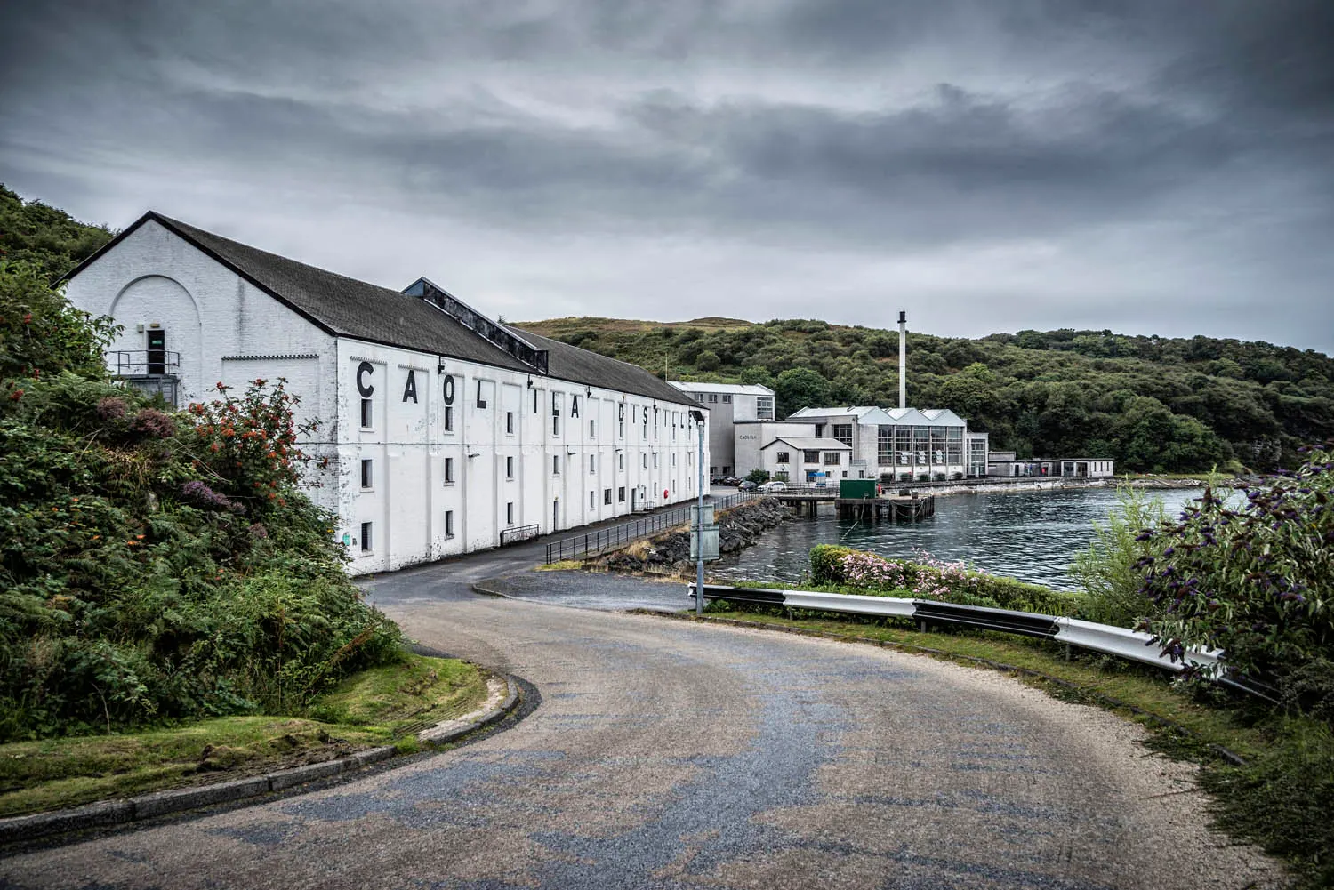 Caol Ila distillery is seen from a winding road which leads to the front of the building where the distillery meets the bay. Behind the distillery are hills with lots of green foliage.