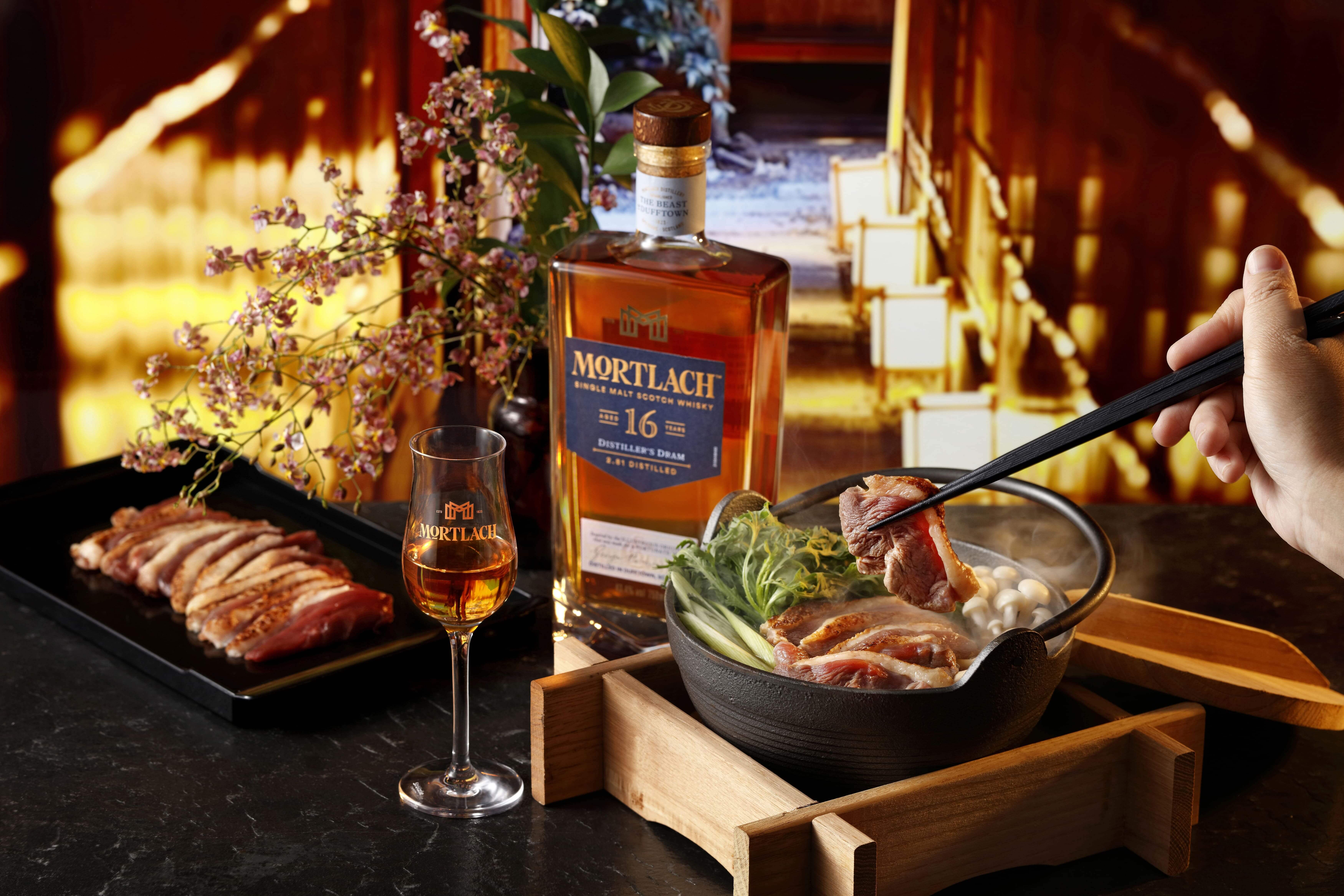 一個人用筷子從火鍋中夾起肉，旁邊有一瓶和一杯Mortlach 16威士忌，切好的肉和裝飾性的花朵放在一張暗色的桌子上。