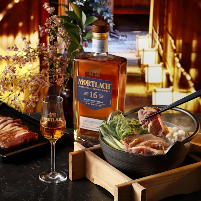 一個人用筷子從火鍋中夾起肉,旁邊有一瓶和一杯Mortlach 16威士忌,切好的肉和裝飾性的花朵放在一張暗色的桌子上。