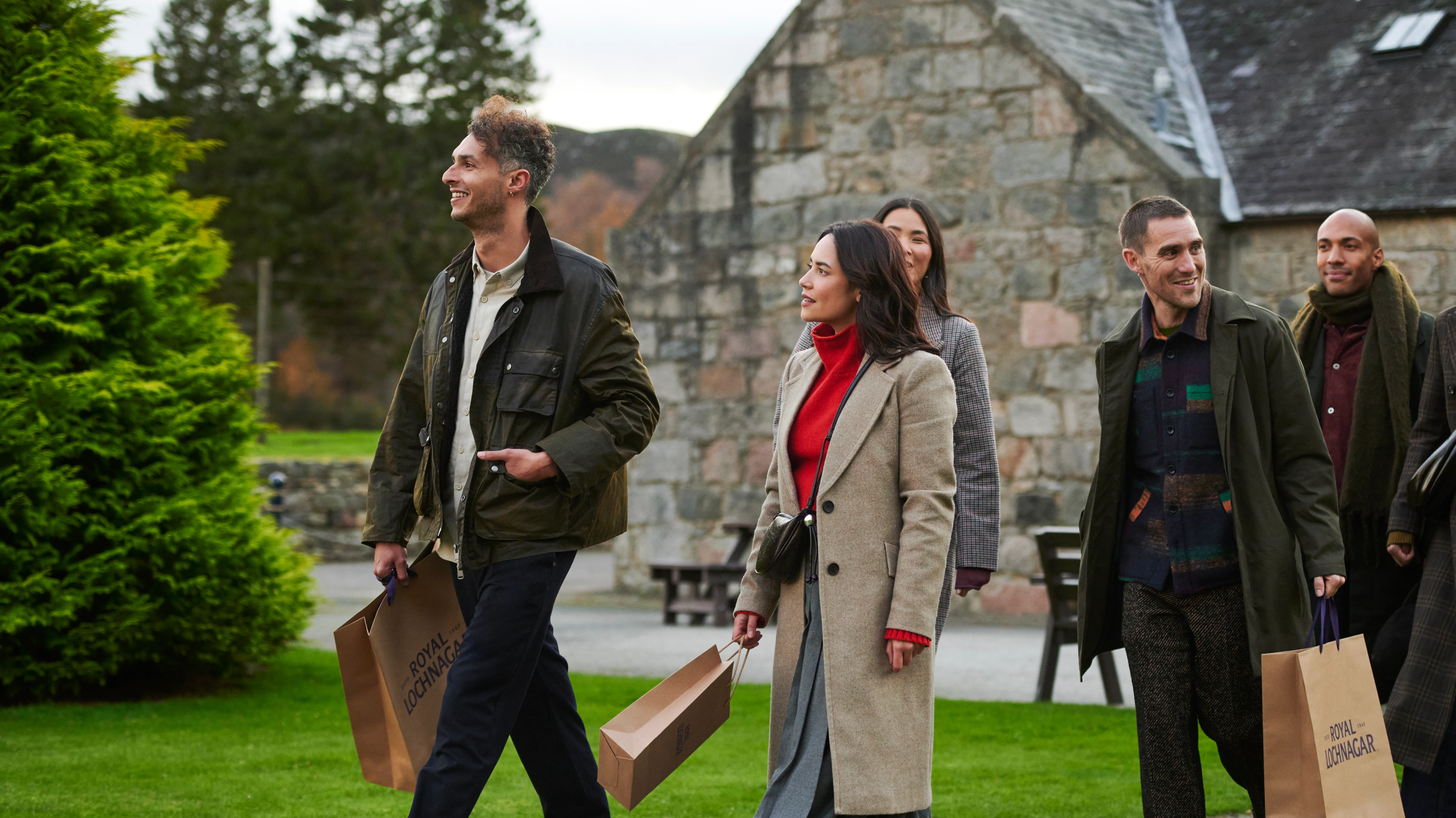 A group of friends leave Royal Lochnagar distillery, walking away from the building and carrying gift bags. They are all smiling.