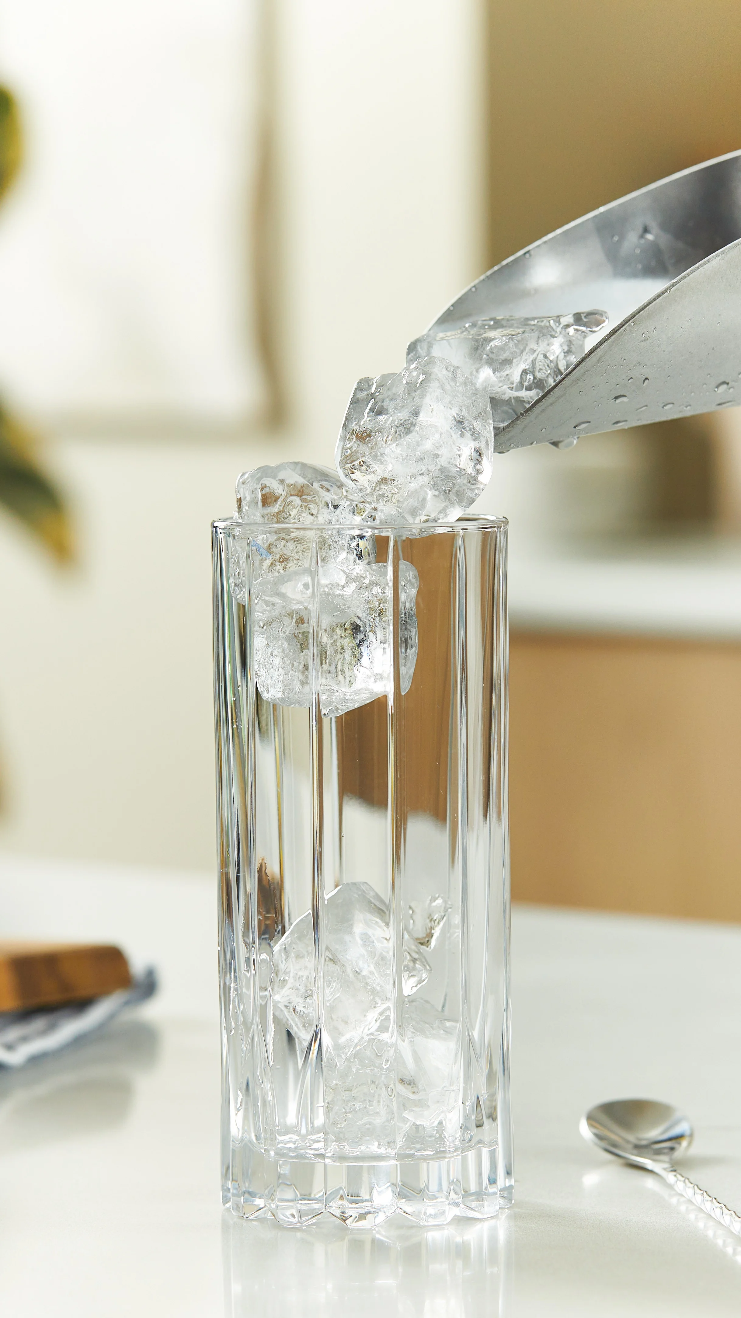 Square ice cubes being poured into a clear highball glass.