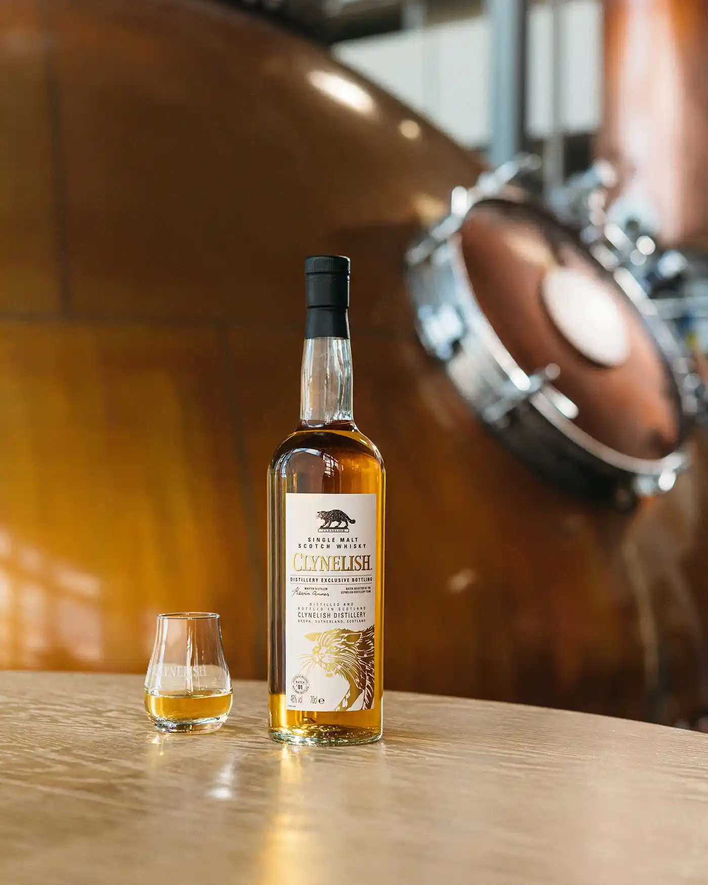 A bottle of Clynelish whisky sits on a wooden tabletop. Behind the bottle is a metal whisky still. Next to the bottle is a dram glass containing whisky.