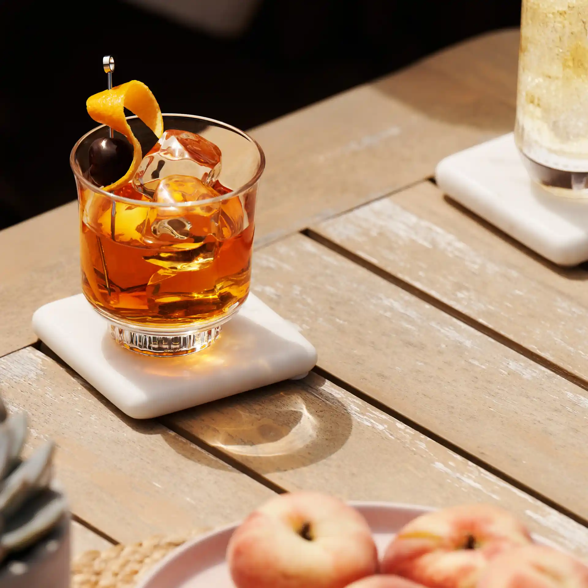 Ein Old-Fashioned-Cocktail liegt in der Sonne auf einer hölzernen Unterlage, daneben stehen eine kleine Pflanze und eine Schale mit Pfirsichen.