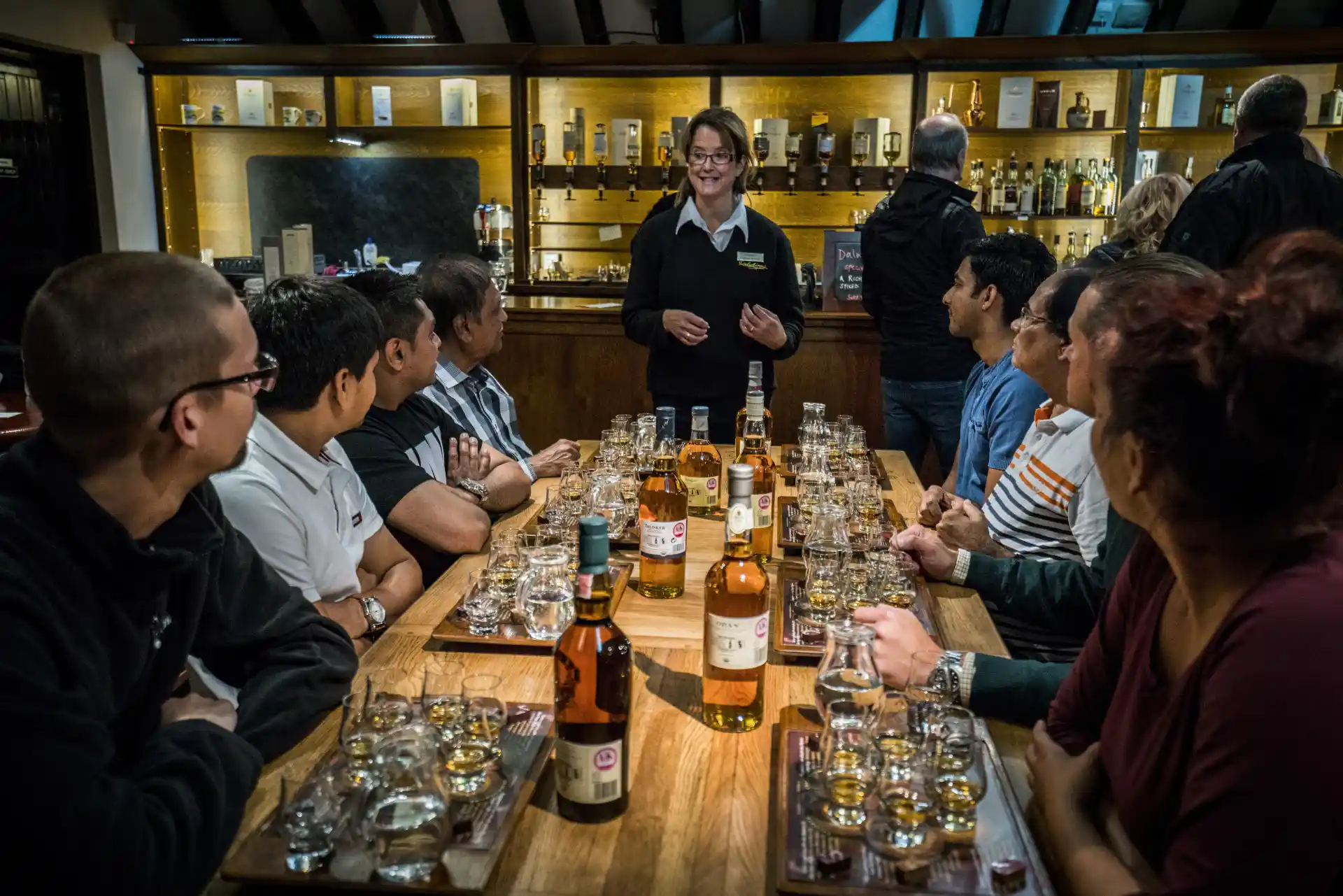 Een expert leidt een proeverij met een groep mensen aan een tafel. Op de tafel staan flessen whisky en dram-glazen. Achter de expert staat een bar waar klanten aan staan, met veel whiskyflessen uitgestald op planken.