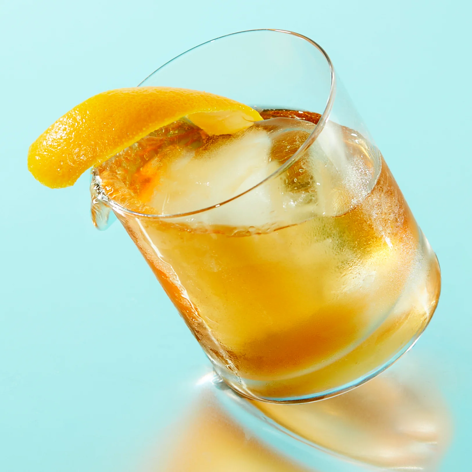 A rocks glass tilted on its side, filled with golden liquid, one large ice cube and an orange garnish.
