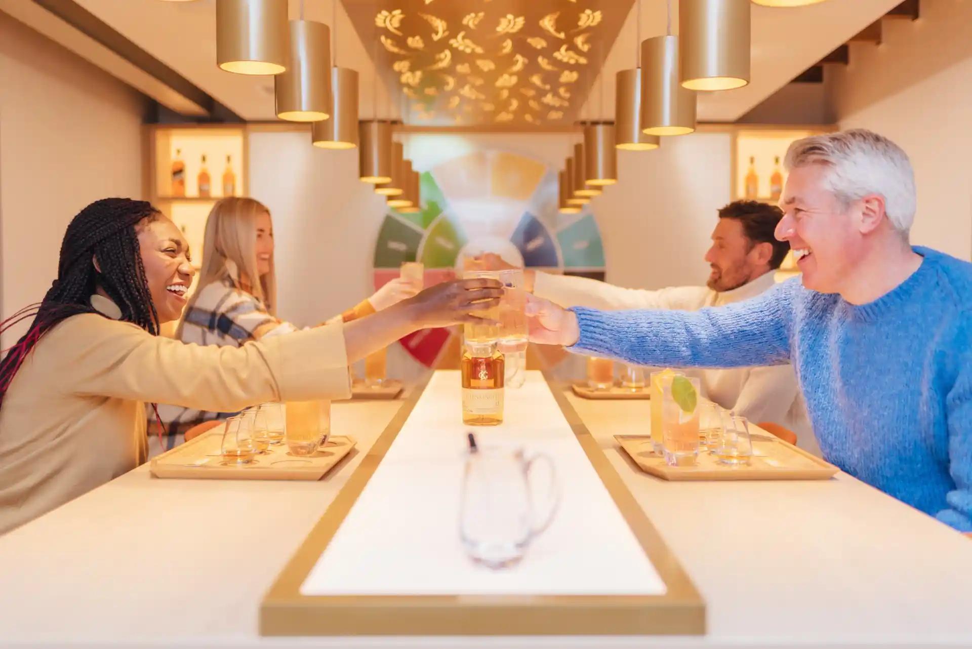 A group of four people sit around a table. They each hold a glass aloft to ‘cheers’. On the table are assorted whisky drams and highball glasses containing whisky and cocktails.