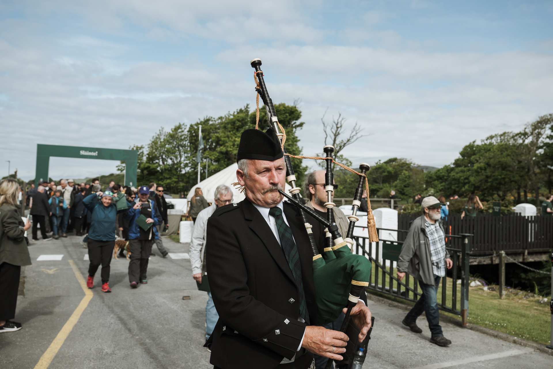 Ein traditionell gekleideter Dudelsackspieler führt eine Parade während des Feis Ile Festivals auf Islay an. Eine Menschenmenge folgt ihm durch einen Eingangsbereich mit der Aufschrift 'Slàinte!'.