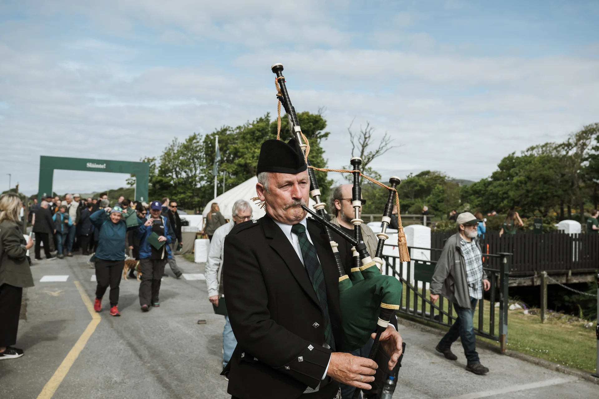 Ein traditionell gekleideter Dudelsackspieler führt eine Parade während des Feis Ile Festivals auf Islay an. Eine Menschenmenge folgt ihm durch einen Eingangsbereich mit der Aufschrift 'Slàinte!'.